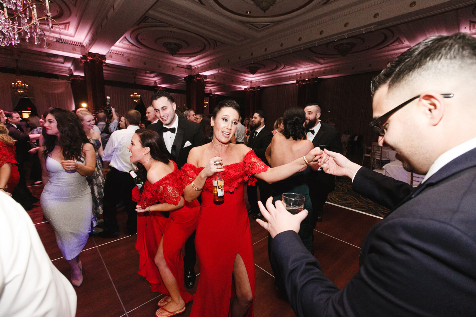 At the elegant Crystal Tea Room wedding, guests in formal attire swirl across the floor. Women in striking red dresses, drink in hand, mingle under ornate ceilings and soft ambient lighting, adding an air of sophistication to the evening.