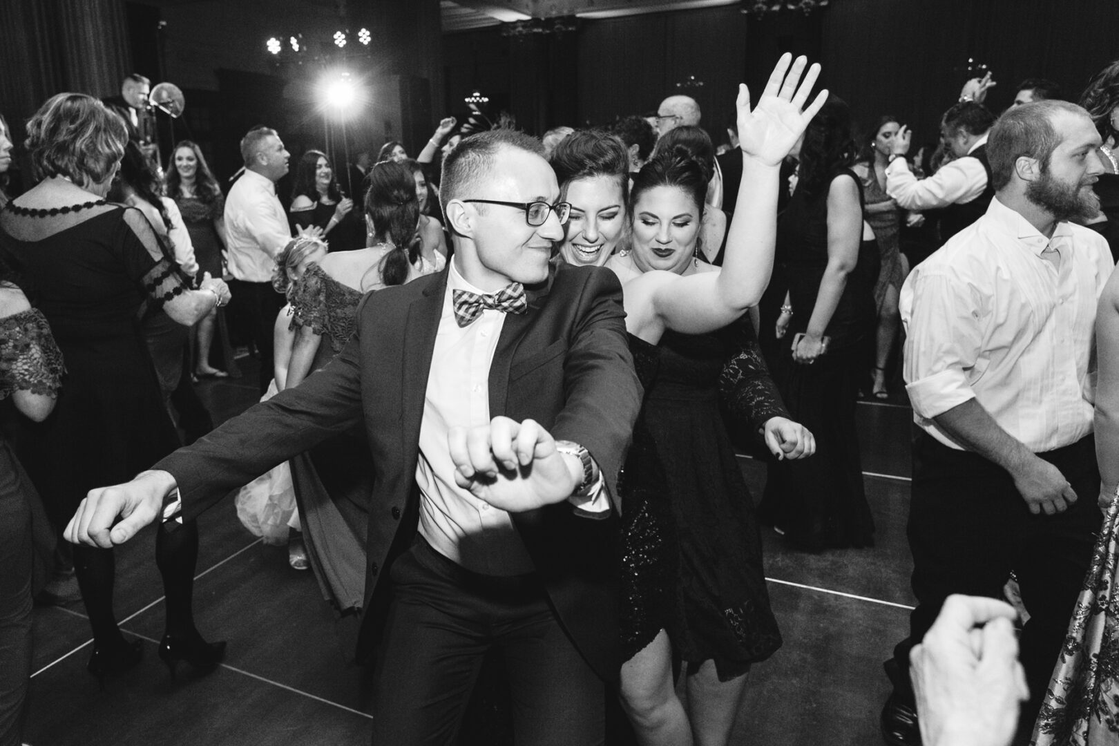 People dancing energetically at an indoor event in the Crystal Tea Room, with others in the background. The scene is lively and festive, reminiscent of a joyous wedding celebration, and everyone seems to be enjoying the moment.