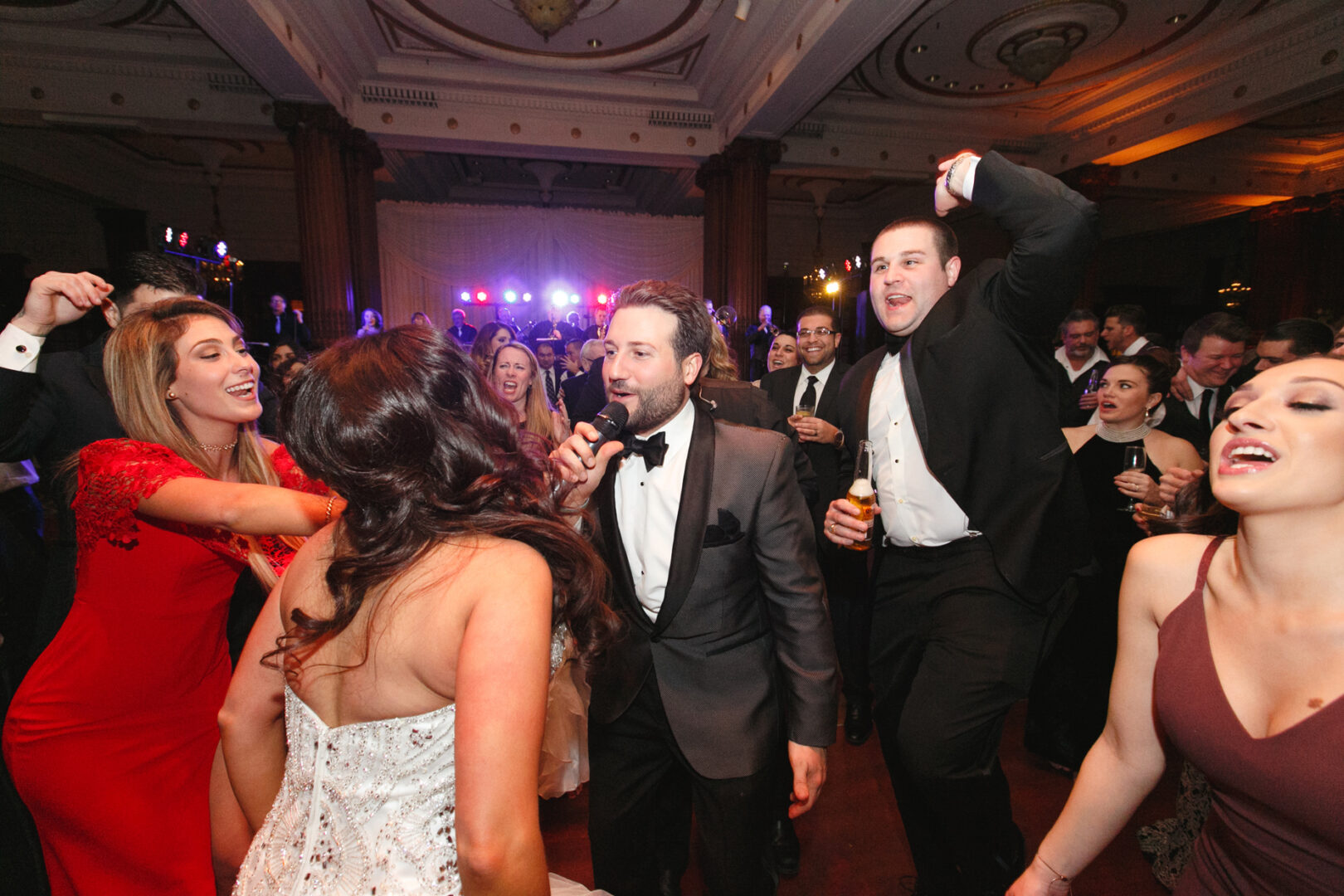 A lively Crystal Tea Room wedding reception unfolds with a group of people dancing and singing. Men in suits and women in elegant dresses revel in the celebration within the beautifully decorated venue, where dim lighting adds a touch of magic.