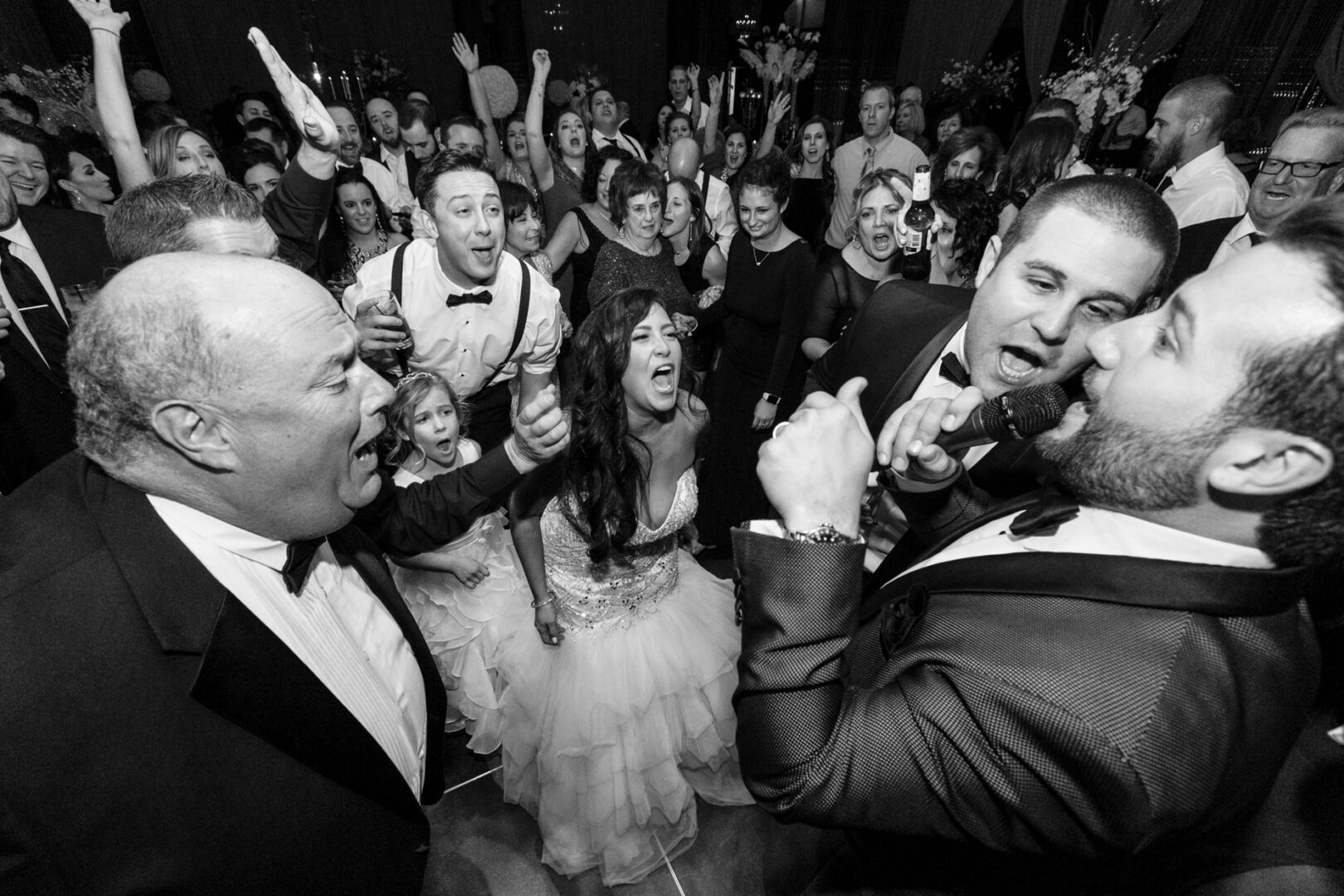 A lively group of people in formal attire energetically singing and cheering, celebrating a glamorous Crystal Tea Room wedding in a crowded indoor setting.
