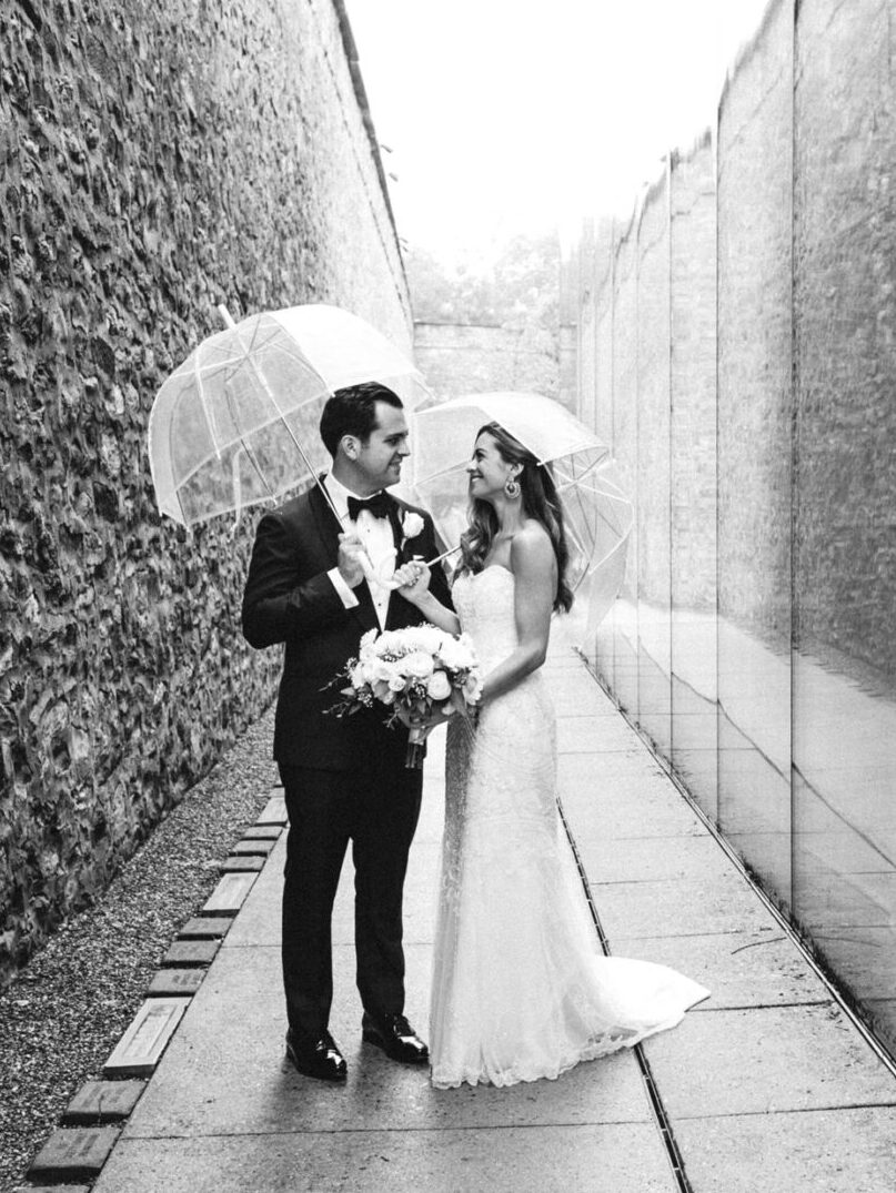 Bride and groom stand under clear umbrellas beside a reflective glass wall at their Michener Art Museum wedding, holding a bouquet, with a stone wall on the opposite side.