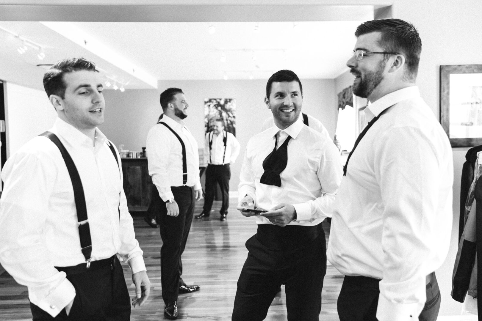 Five men in dress shirts and suspenders stand in a room at the Michener Art Museum, talking and smiling. One holds a phone, capturing the joyous moments of what appears to be a wedding day. The room’s wooden floors and large window add a touch of elegance to the setting.