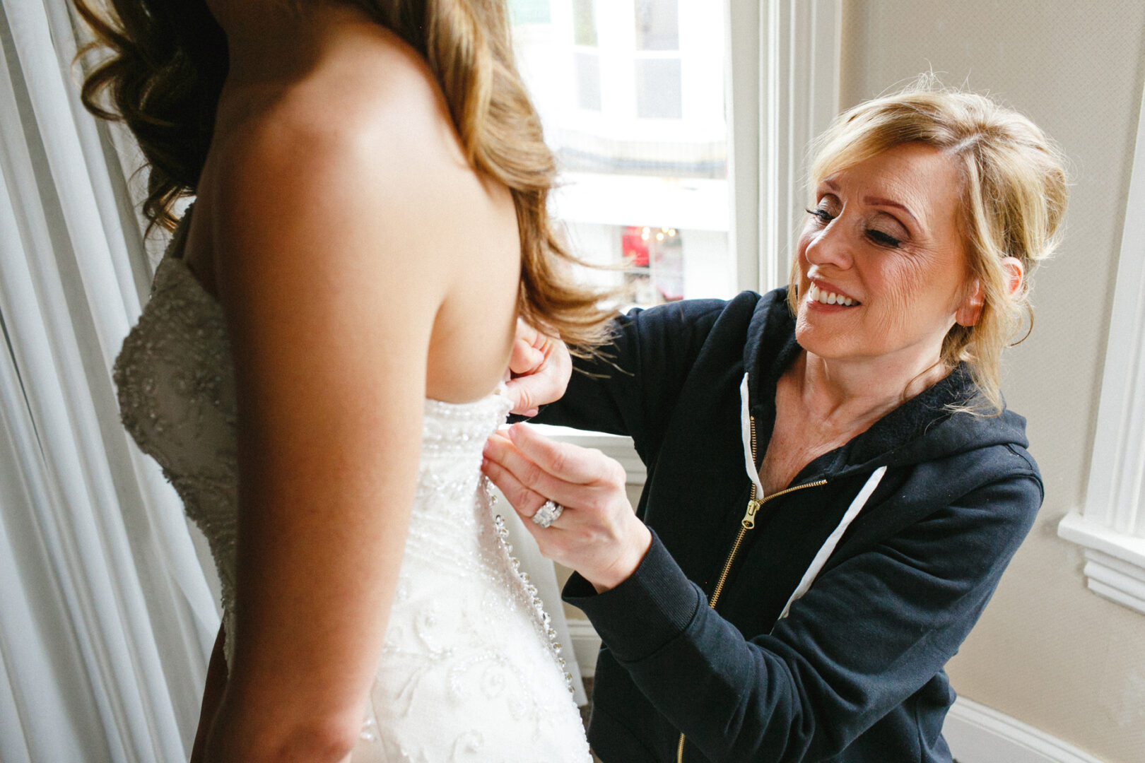 At the Michener Art Museum, a woman in a dark jacket assists another in adjusting her white wedding dress near a window.