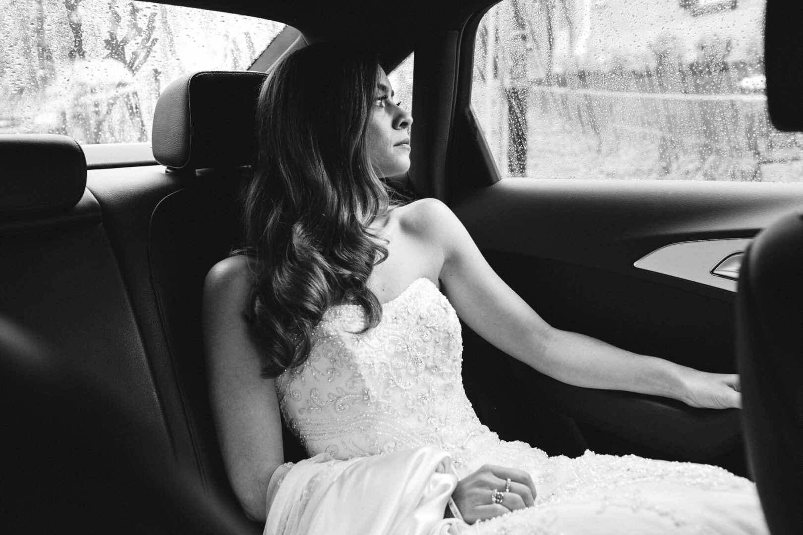 In a strapless dress, the bride sits in the backseat of a car, gazing wistfully out of a rain-specked window, her thoughts drifting to the magical Michener Art Museum wedding that awaits.