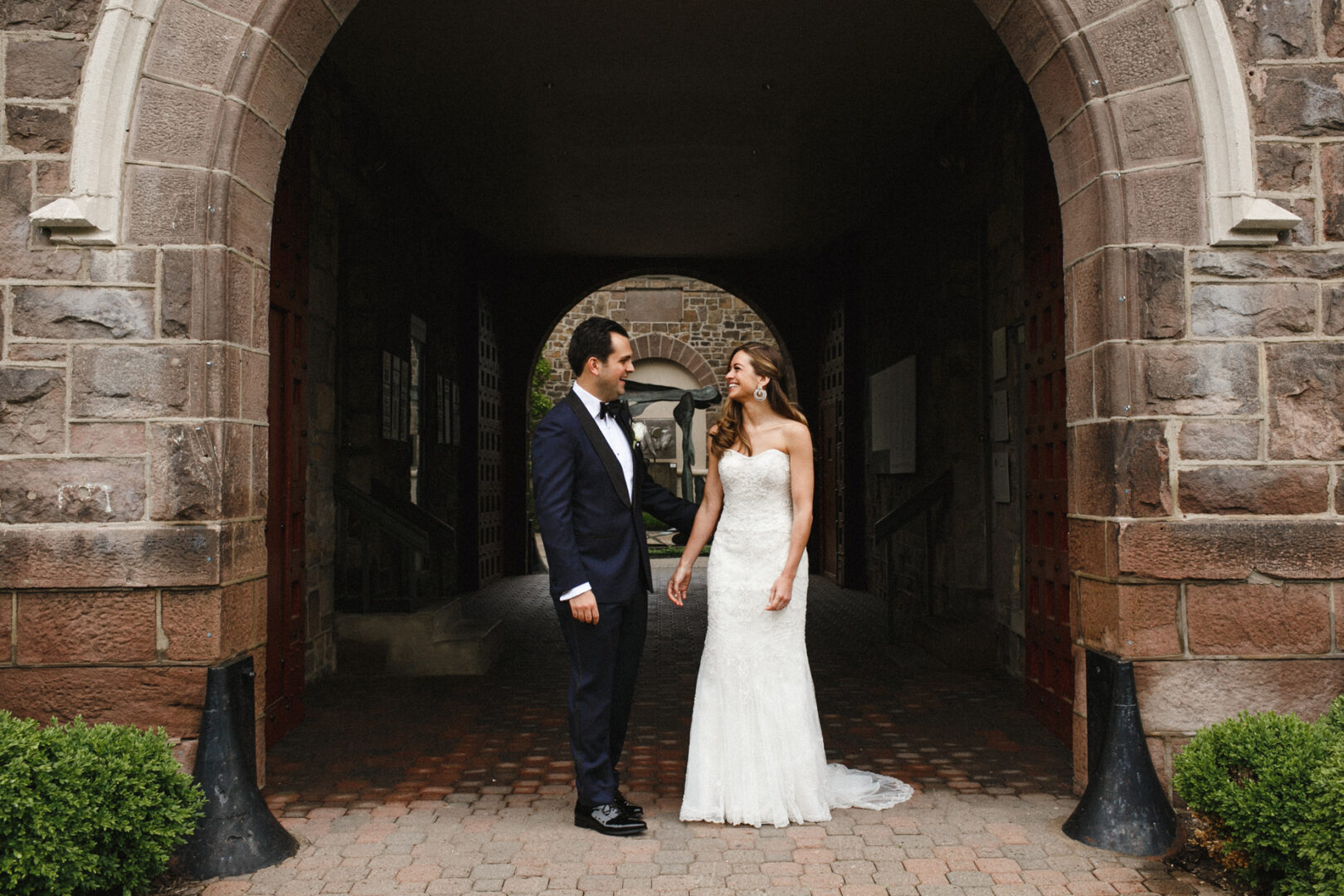 A bride in a white gown and a groom in a dark suit stand facing each other under the stone archway at the picturesque Michener Art Museum, creating an unforgettable wedding backdrop.
