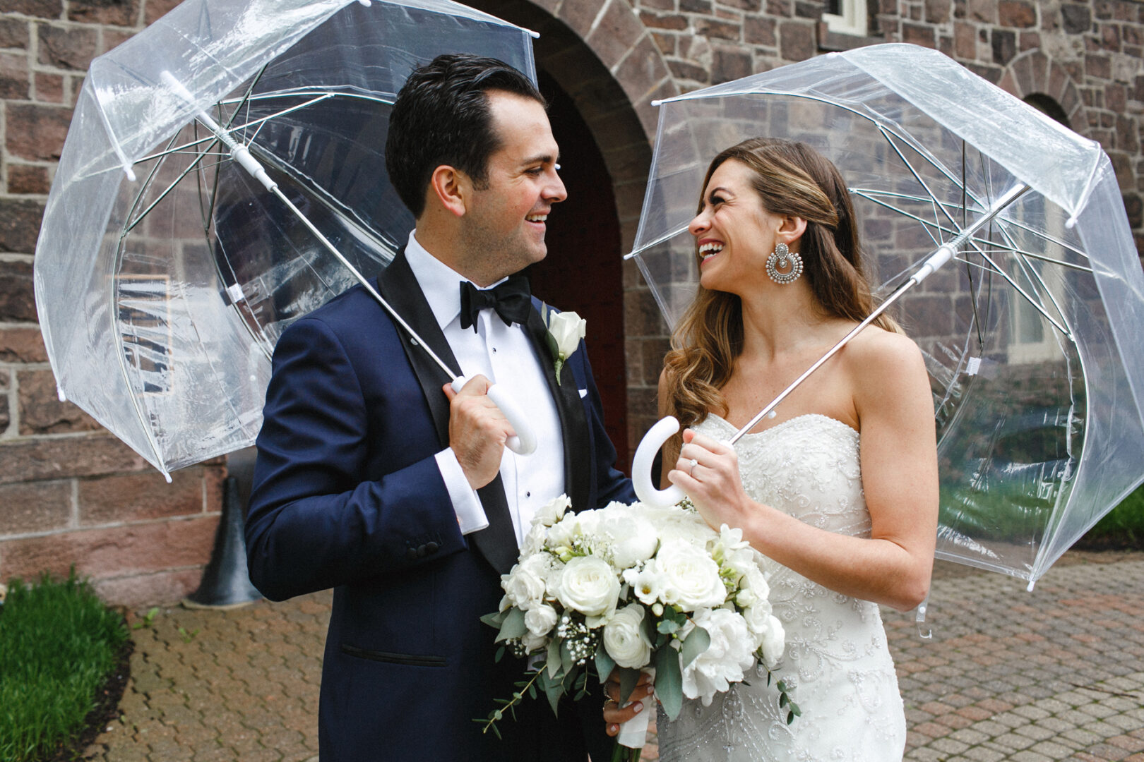 A bride and groom smile at each other under clear umbrellas, the bride clutching a bouquet of white flowers as they stand in front of the stone façade of the Michener Art Museum, capturing an enchanting wedding moment.
