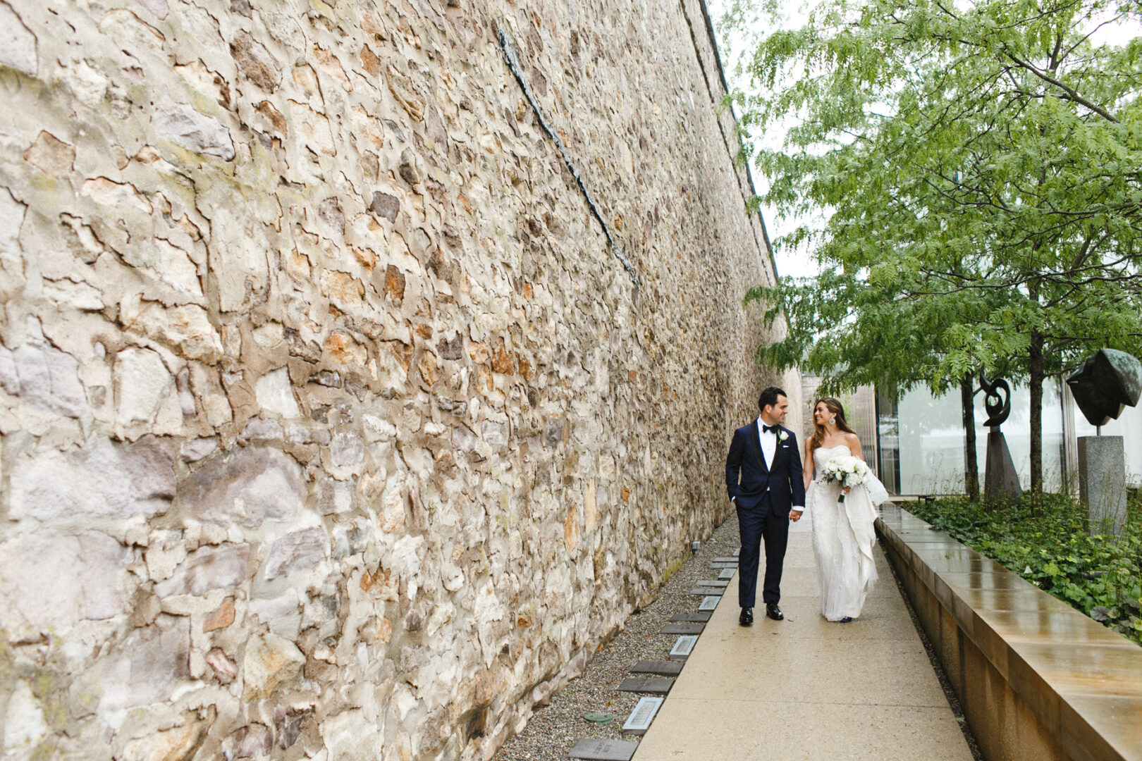A bride and groom walk hand in hand along a path beside a stone wall and lush greenery at the Michener Art Museum Wedding.