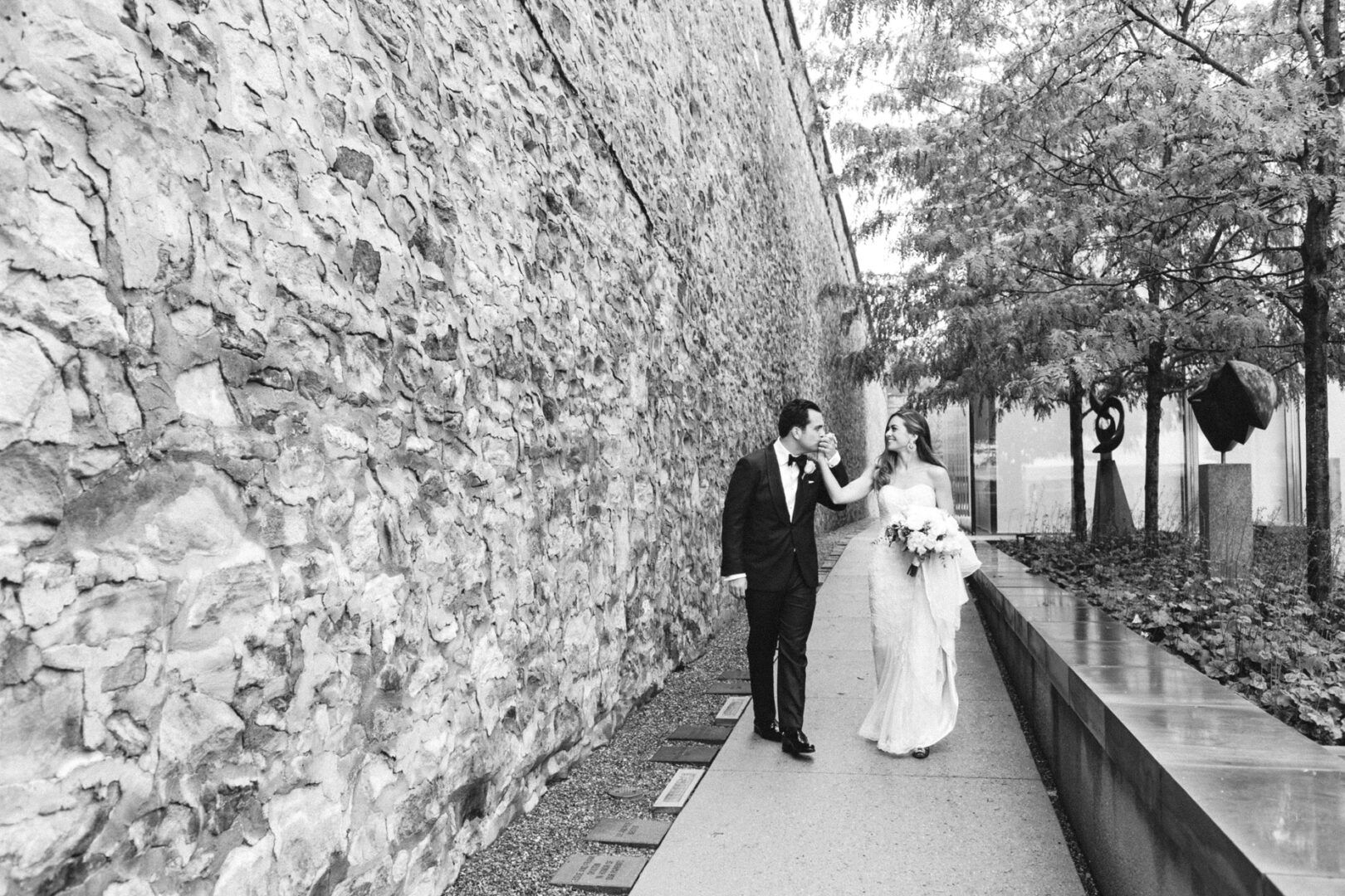 At the Michener Art Museum wedding, a bride and groom grace the stone wall path, with the groom tenderly kissing her hand. She clutches a bouquet, as trees elegantly line their journey on the right.