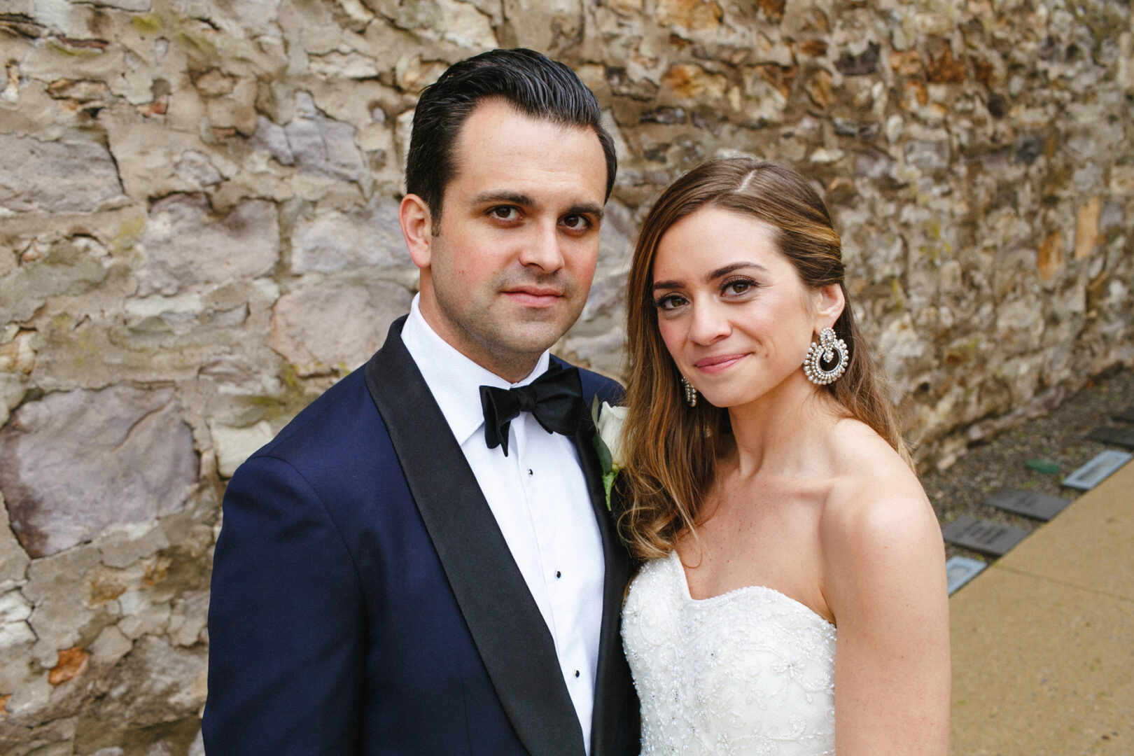 A couple in formal wedding attire poses gracefully in front of the historic stone wall at the Michener Art Museum, capturing an exquisite moment from their special day.