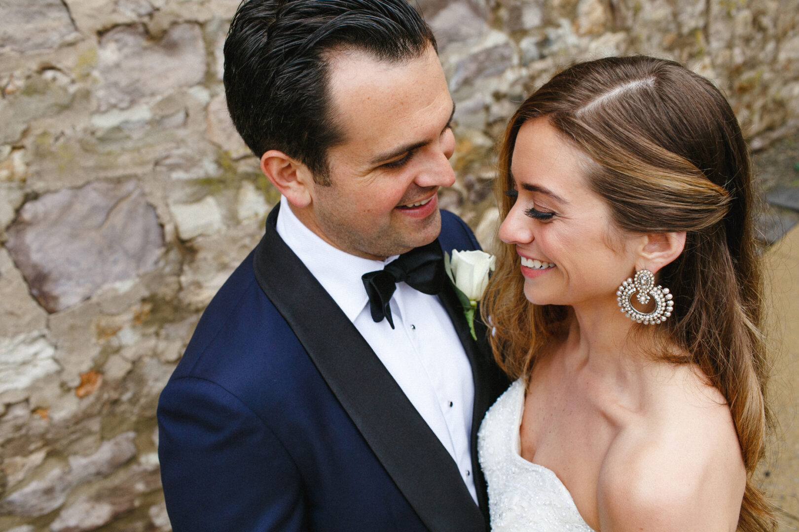 A couple wearing formal attire stands close, smiling at each other in front of a stone wall, capturing the essence of their Michener Art Museum wedding.