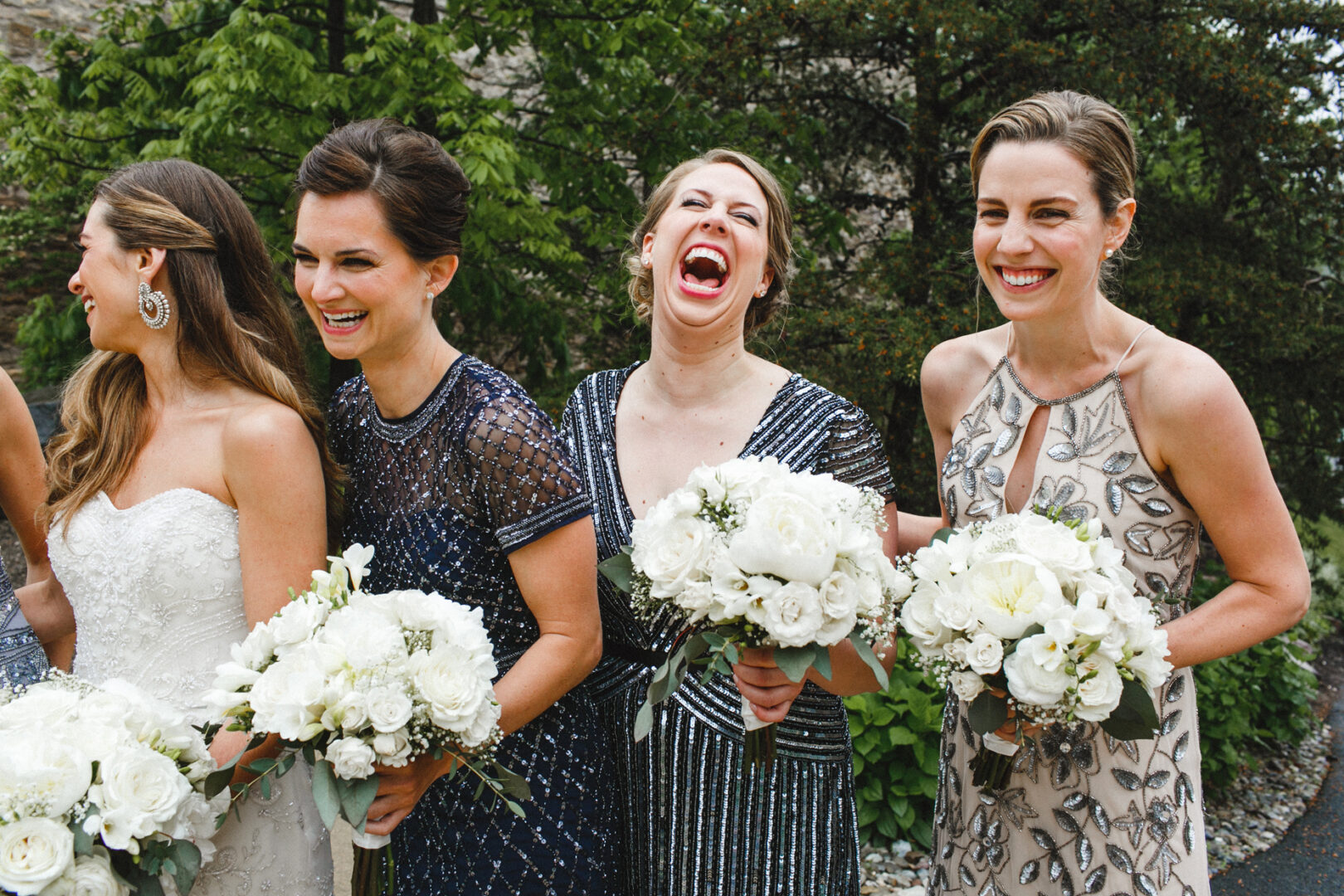 Four women in dresses laugh while holding white bouquets, standing outdoors amidst the scenic backdrop of the Michener Art Museum’s lush gardens.
