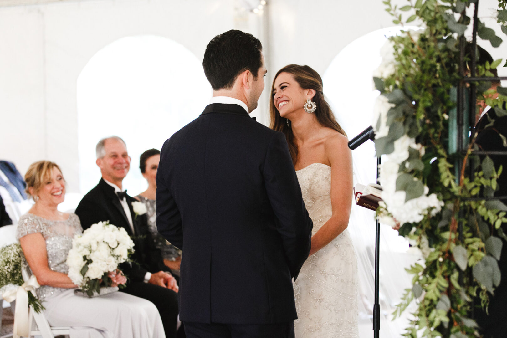 A bride and groom stand facing each other during a Michener Art Museum wedding ceremony. Guests are seated nearby, with one holding a bouquet of white flowers. Lush greenery decorates the space, adding to the artistic ambiance.