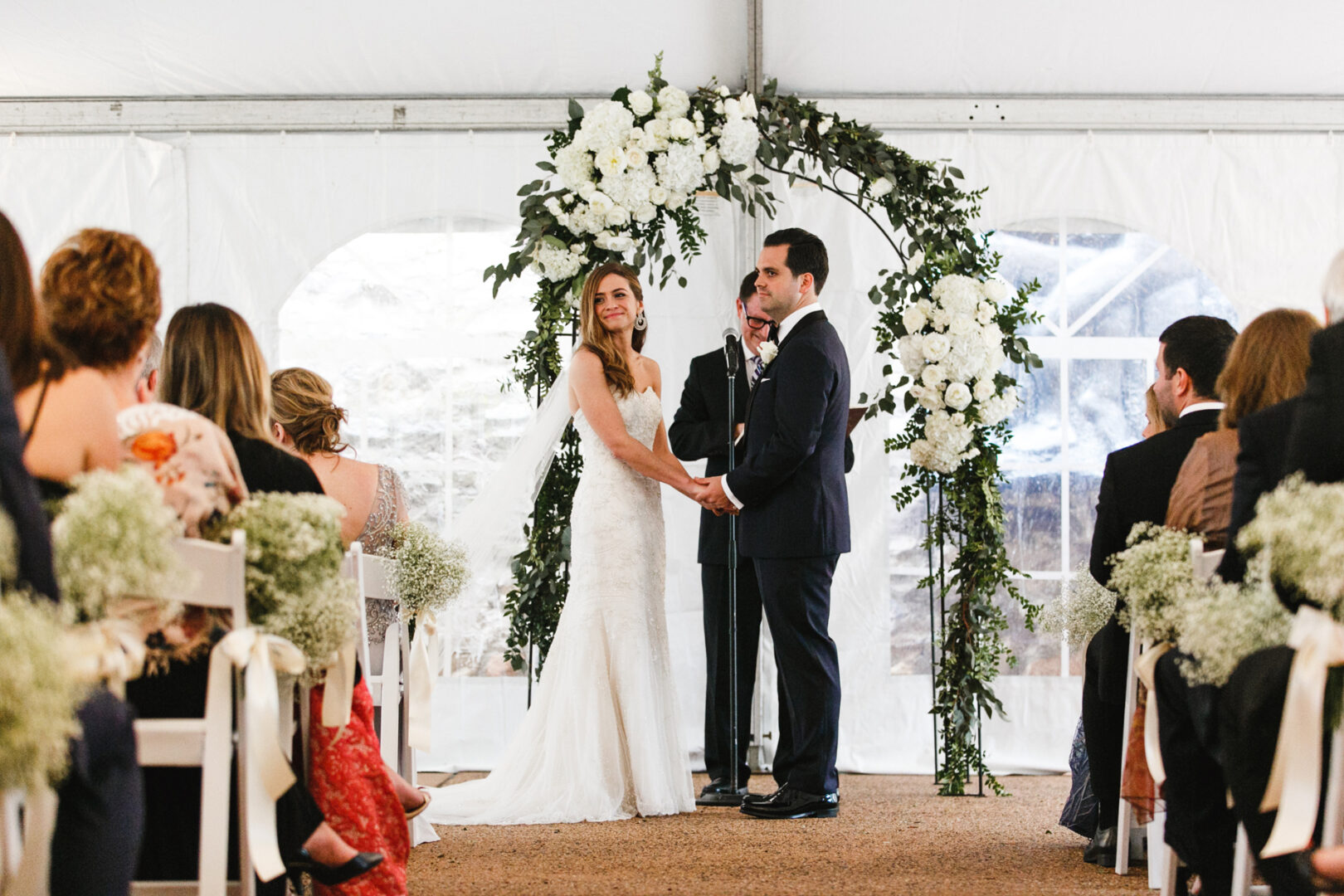 The bride and groom exchange vows under a floral arch at their Michener Art Museum wedding, surrounded by seated guests inside a beautifully draped tent.