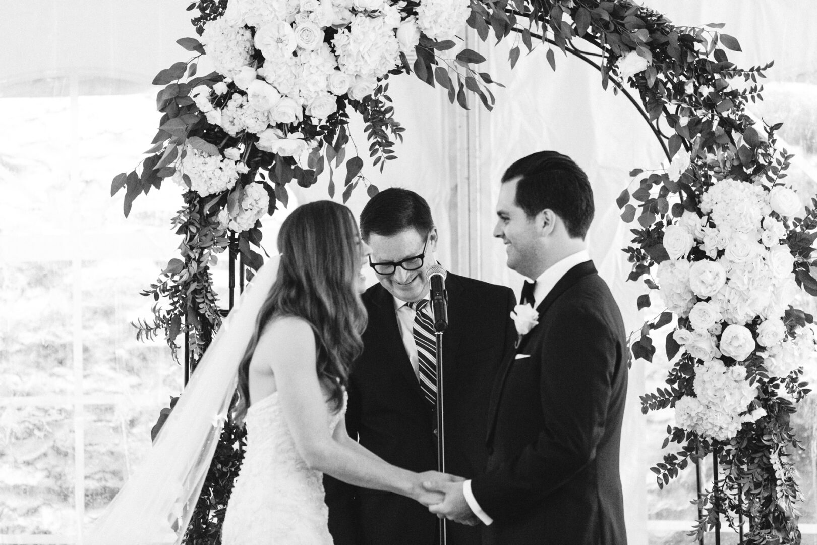 A bride and groom hold hands and smile under a floral arch at their Michener Art Museum wedding ceremony, officiated by a man with glasses.