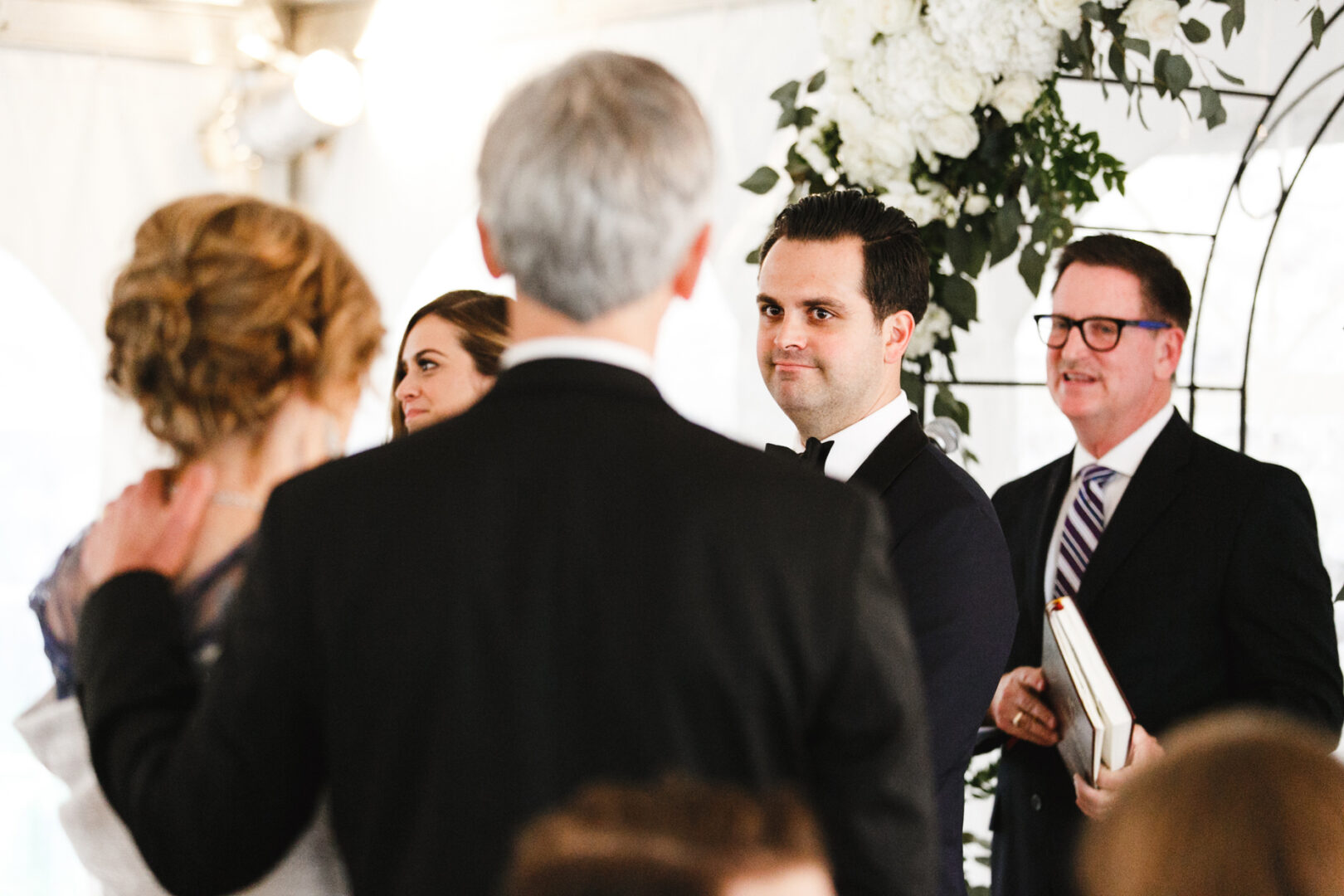 At a Michener Art Museum wedding, the groom gazes at the couple while an officiant with a book stands nearby. Exquisite floral arrangements add elegance to this beautiful ceremony.