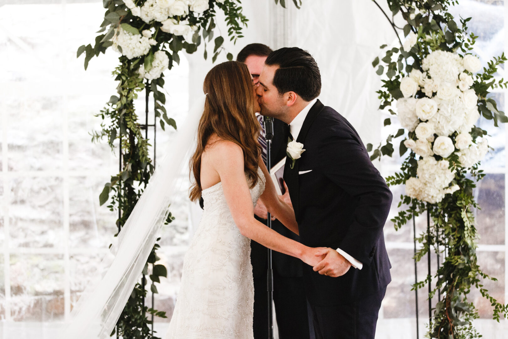 A bride and groom share a tender kiss at their Michener Art Museum wedding, standing under a floral archway adorned with white flowers and greenery.