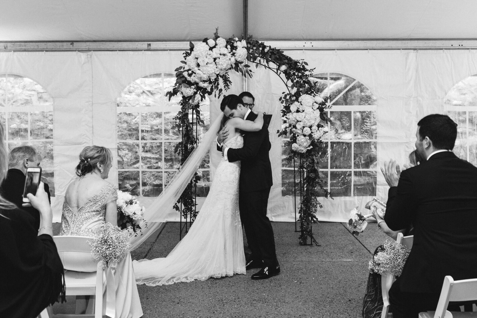 Bride and groom embrace under a floral arch at their Michener Art Museum wedding ceremony, with seated guests watching and capturing the moment.