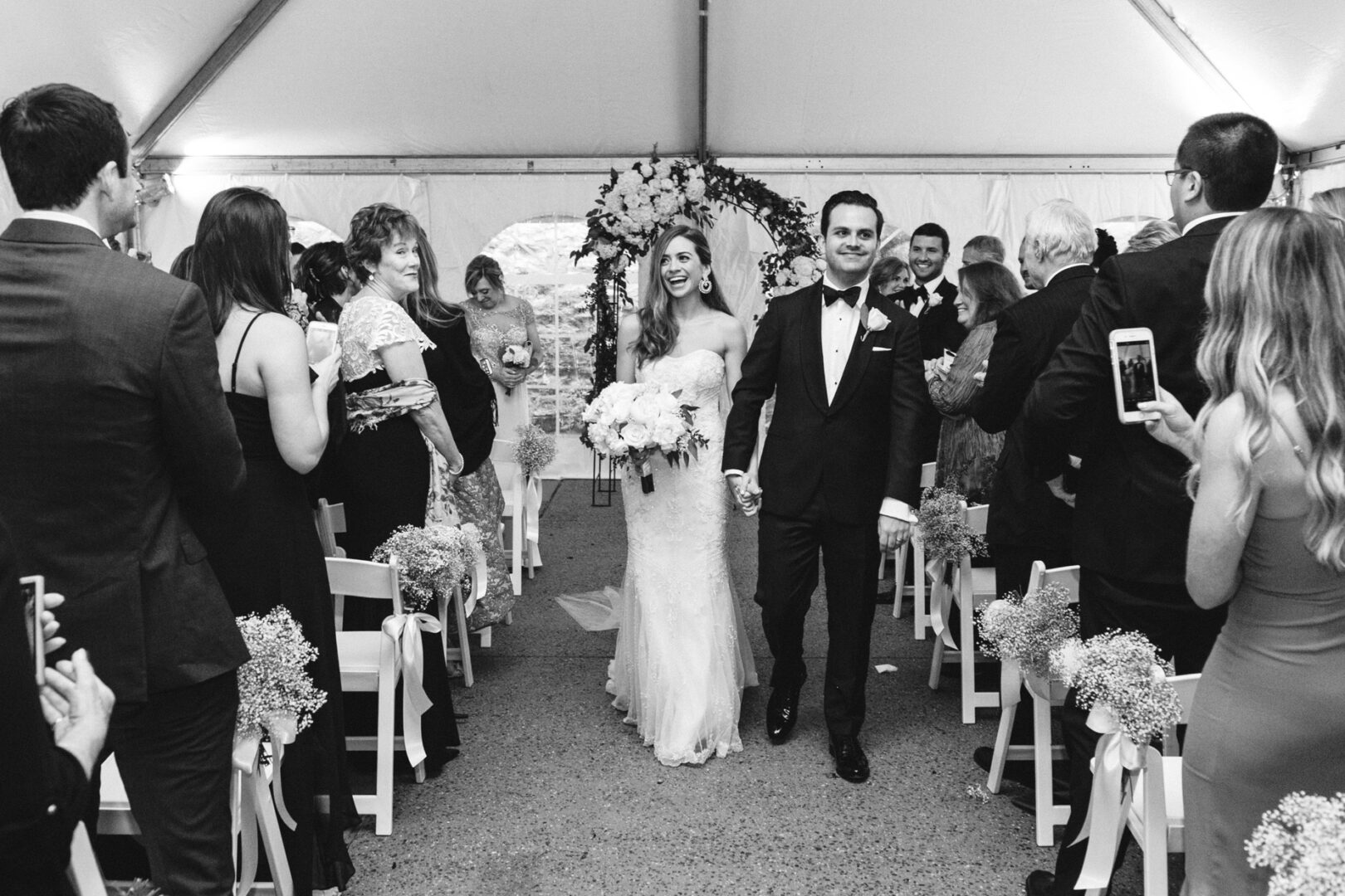 The bride and groom glide down the aisle in a beautifully decorated tent at the Michener Art Museum Wedding, surrounded by guests capturing moments and clapping joyfully.