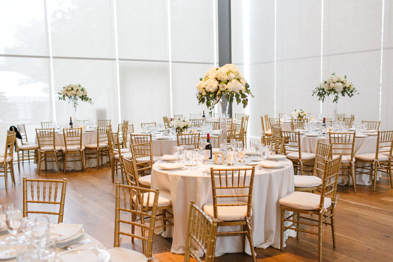Elegant event hall at the Michener Art Museum with round tables covered in white tablecloths, set with glassware and floral centerpieces. Gold chairs surround the tables, and tall arrangements accent the room, perfect for a sophisticated wedding celebration.