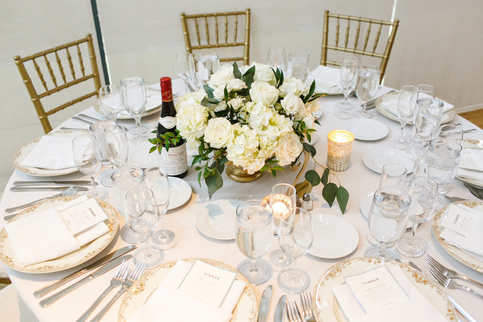 Elegant dining table set with white flowers at the center evokes the sophistication of a Michener Art Museum wedding. The scene is completed by empty glasses, plates, folded napkins, a wine bottle, and candle. Gold-colored chairs and cutlery flank the table.