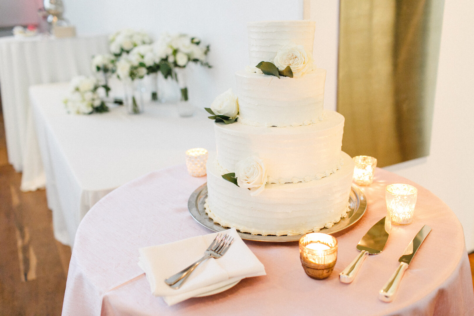 A three-tier white cake adorned with roses graces the table at a Michener Art Museum wedding, accompanied by candles, a knife, and a fork. In the background blooms a bouquet of white flowers.