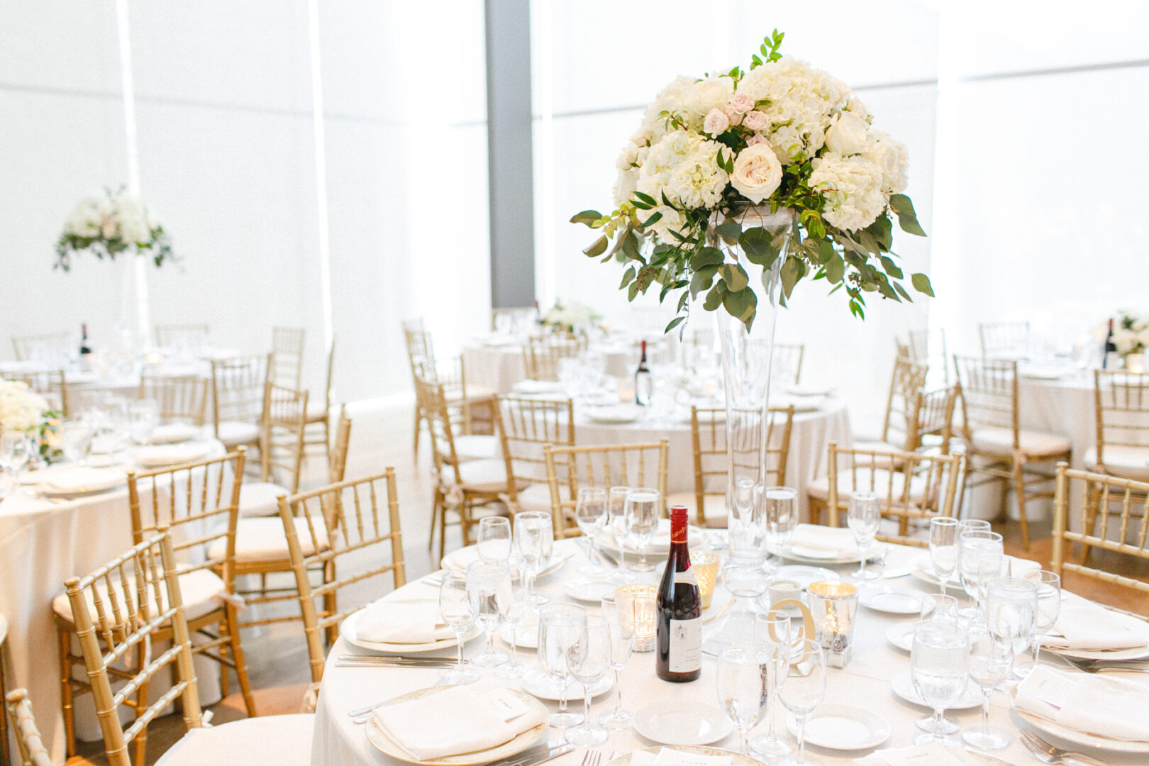 The elegant Michener Art Museum wedding reception features round tables adorned with white tablecloths and floral centerpieces. Wine bottles and glasses gracefully complement gold chairs under the room's natural light.