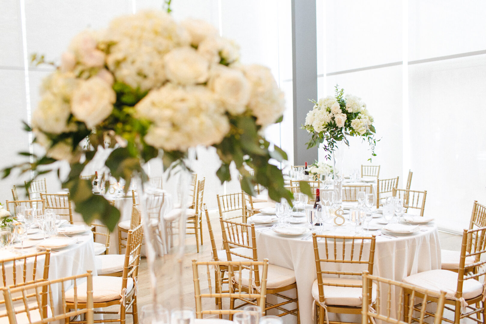 An elegant Michener Art Museum wedding reception features round tables adorned with white tablecloths, beige chairs, and tall floral centerpieces showcasing beautiful white and soft pink flowers.