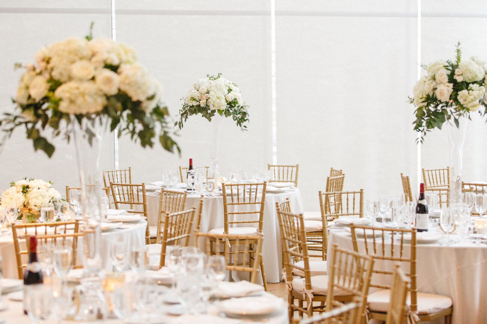 The Michener Art Museum Wedding reception hall features round tables adorned with white linens, gold chairs, and tall floral centerpieces brimming with white flowers and greenery. Elegant glassware and wine bottles are artfully arranged, enveloping guests in an atmosphere of refined celebration.