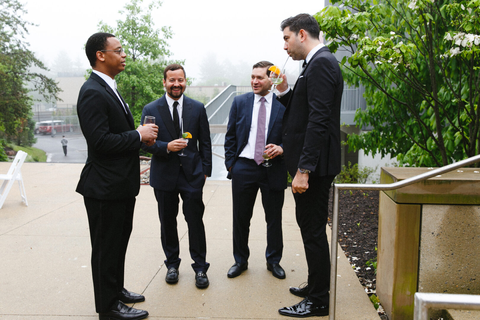 Four men in suits stand outside on a foggy day at the Michener Art Museum wedding, holding drinks and conversing near a staircase and greenery.