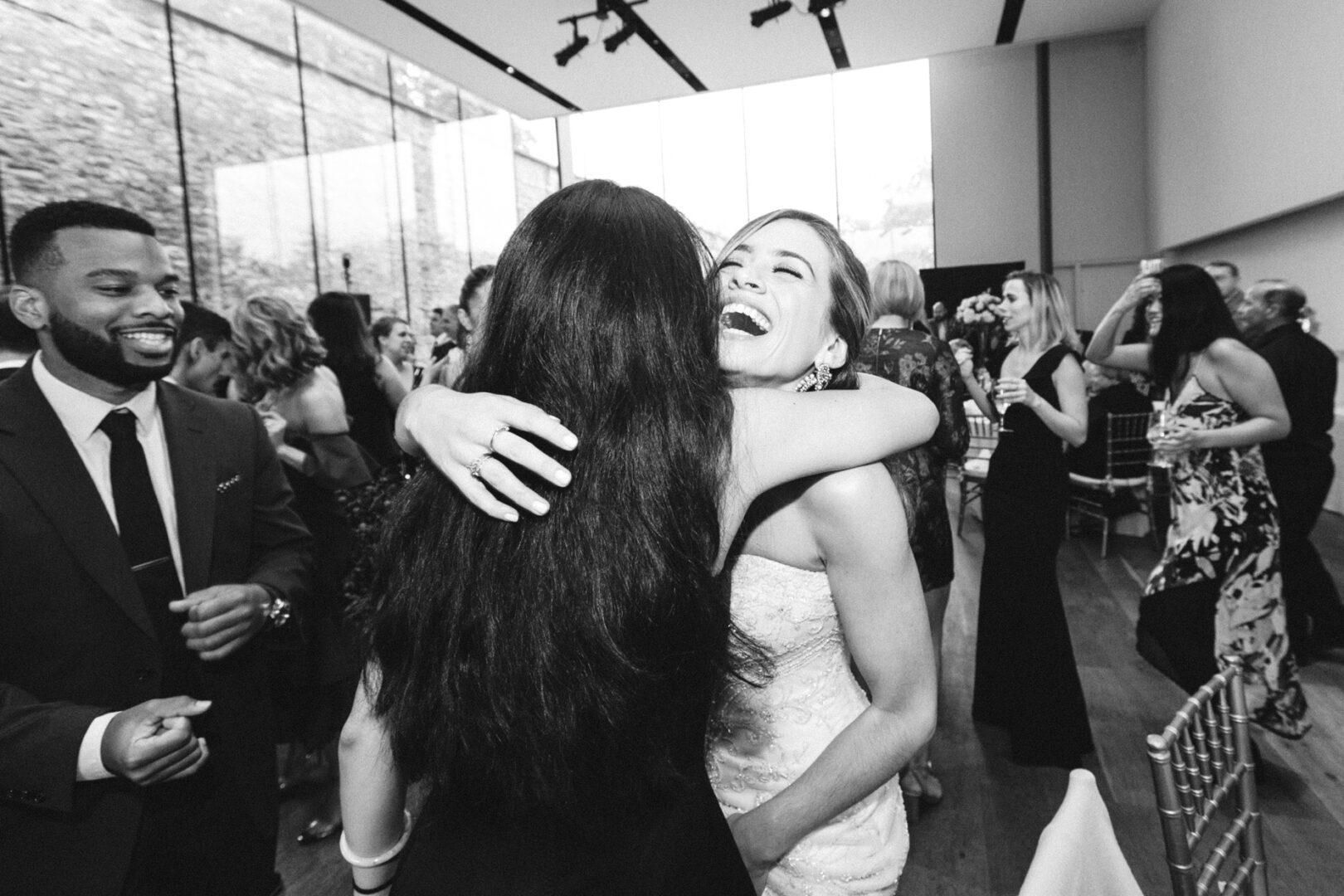 Two women embrace joyfully in a large room at the Michener Art Museum, filled with guests dressed in formal attire. A man in a suit stands nearby, smiling warmly amid the wedding celebration.