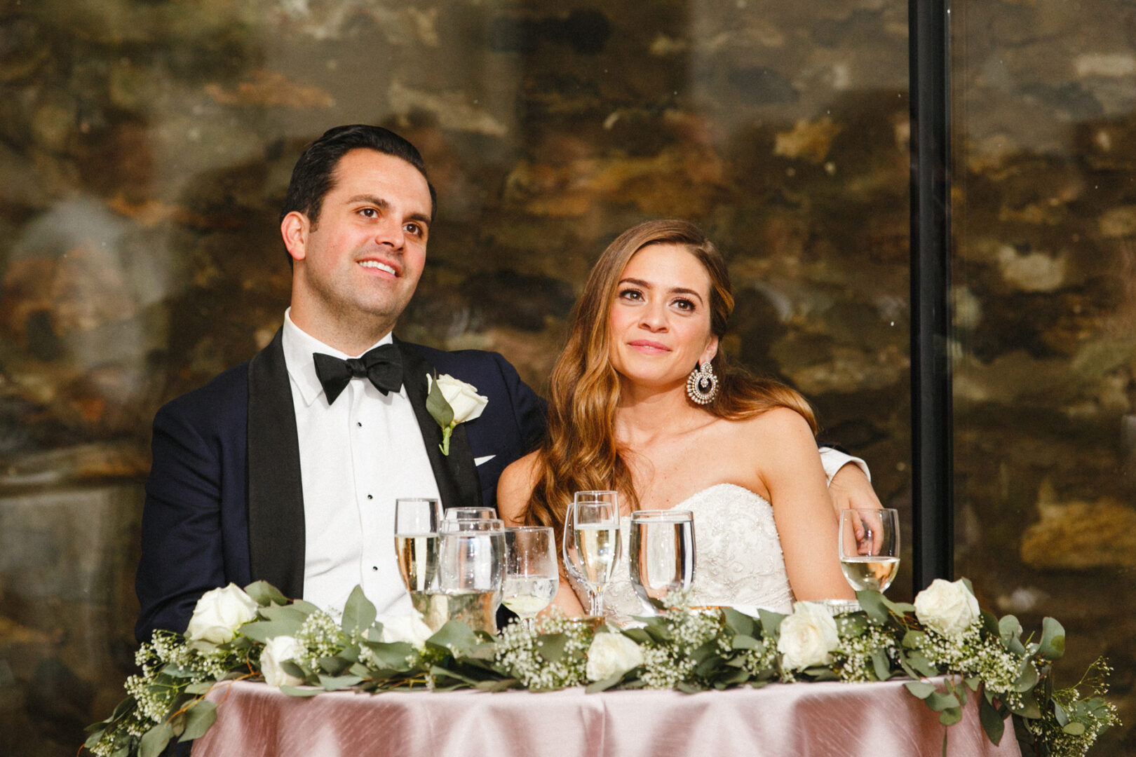 At a table adorned with white roses and greenery, a couple dressed formally enjoys the elegance of a Michener Art Museum wedding reception.
