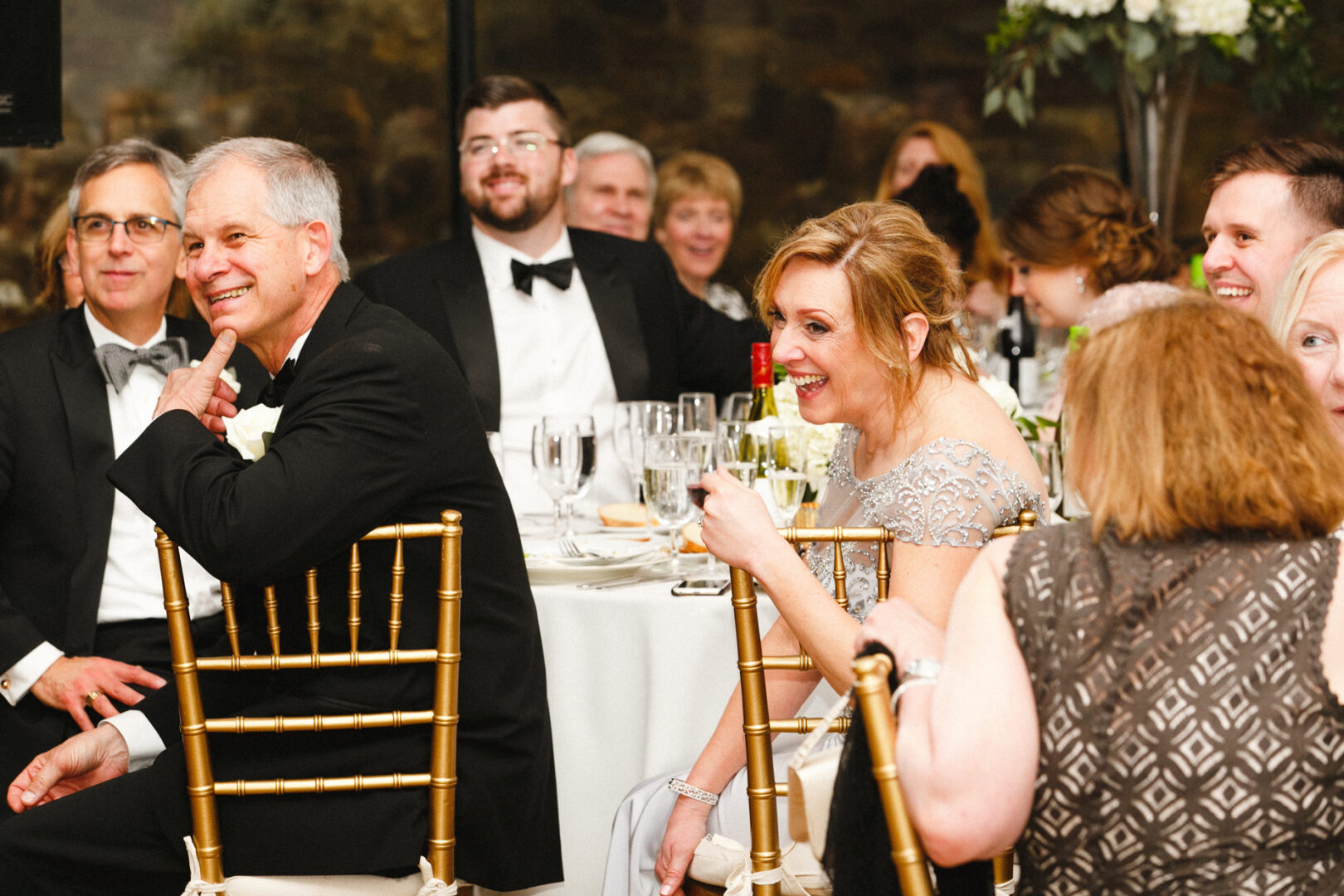 Guests seated at a Michener Art Museum wedding, laughing and engaging with each other. Tables are set with glasses and plates, as several attendees wear elegant suits and formal dresses, adding to the charming ambiance of the event.