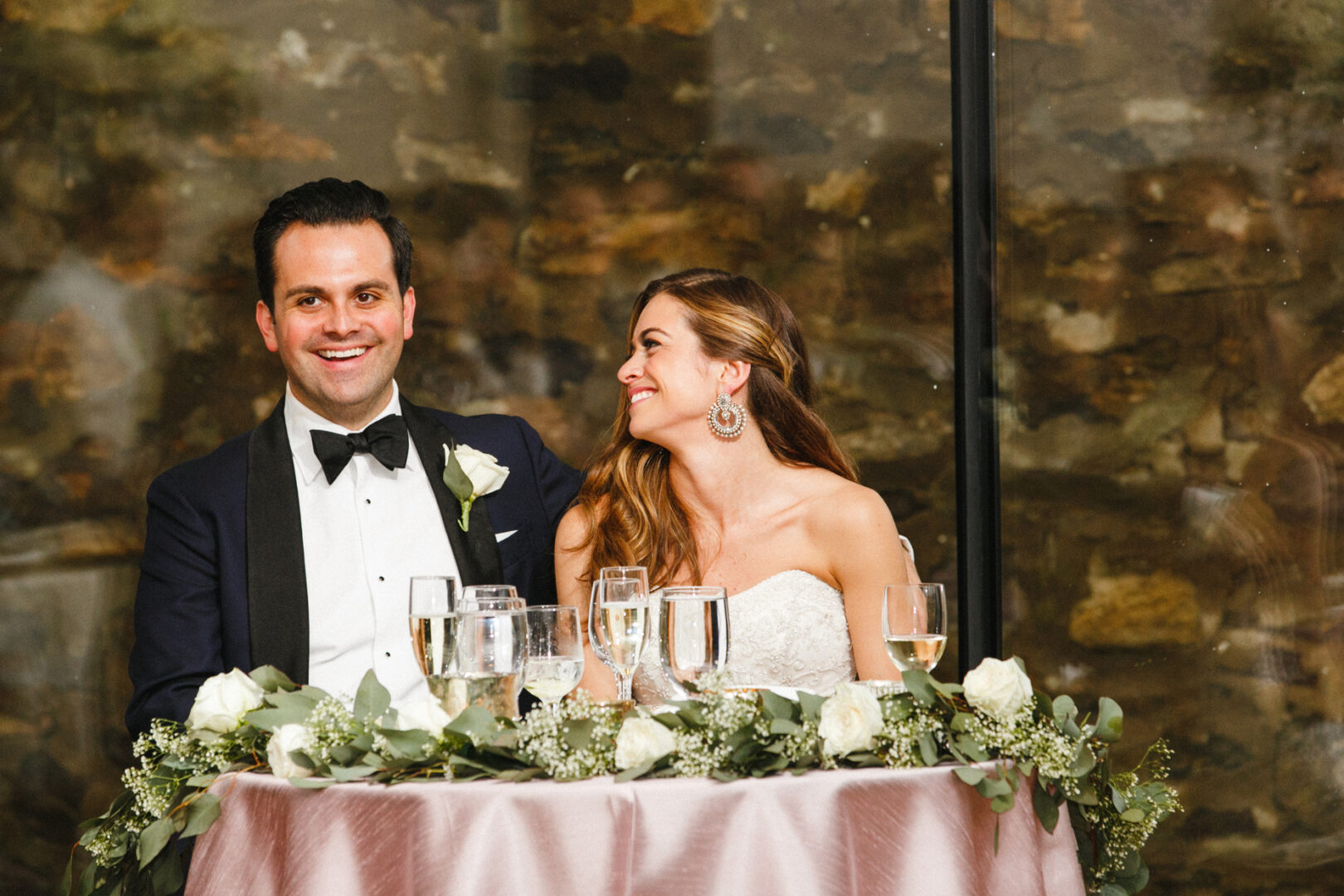 At a table adorned with floral decorations and glasses, a couple in formal attire graces the scene at their Michener Art Museum wedding. The woman gazes at the smiling man, encapsulating a moment of elegance and joy.