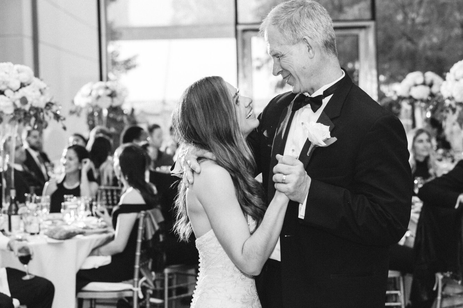 A bride and an older man, both in formal attire, share a dance at the Michener Art Museum wedding reception. Guests and elegantly set tables are visible in the background.
