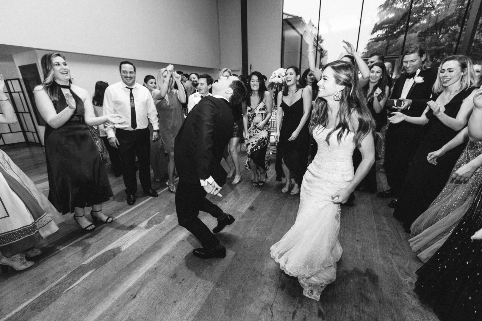 Bride and groom dancing joyfully at their Michener Art Museum wedding, surrounded by guests clapping and smiling in an elegant indoor setting. Captured in timeless black and white, this moment is filled with happiness and celebration.