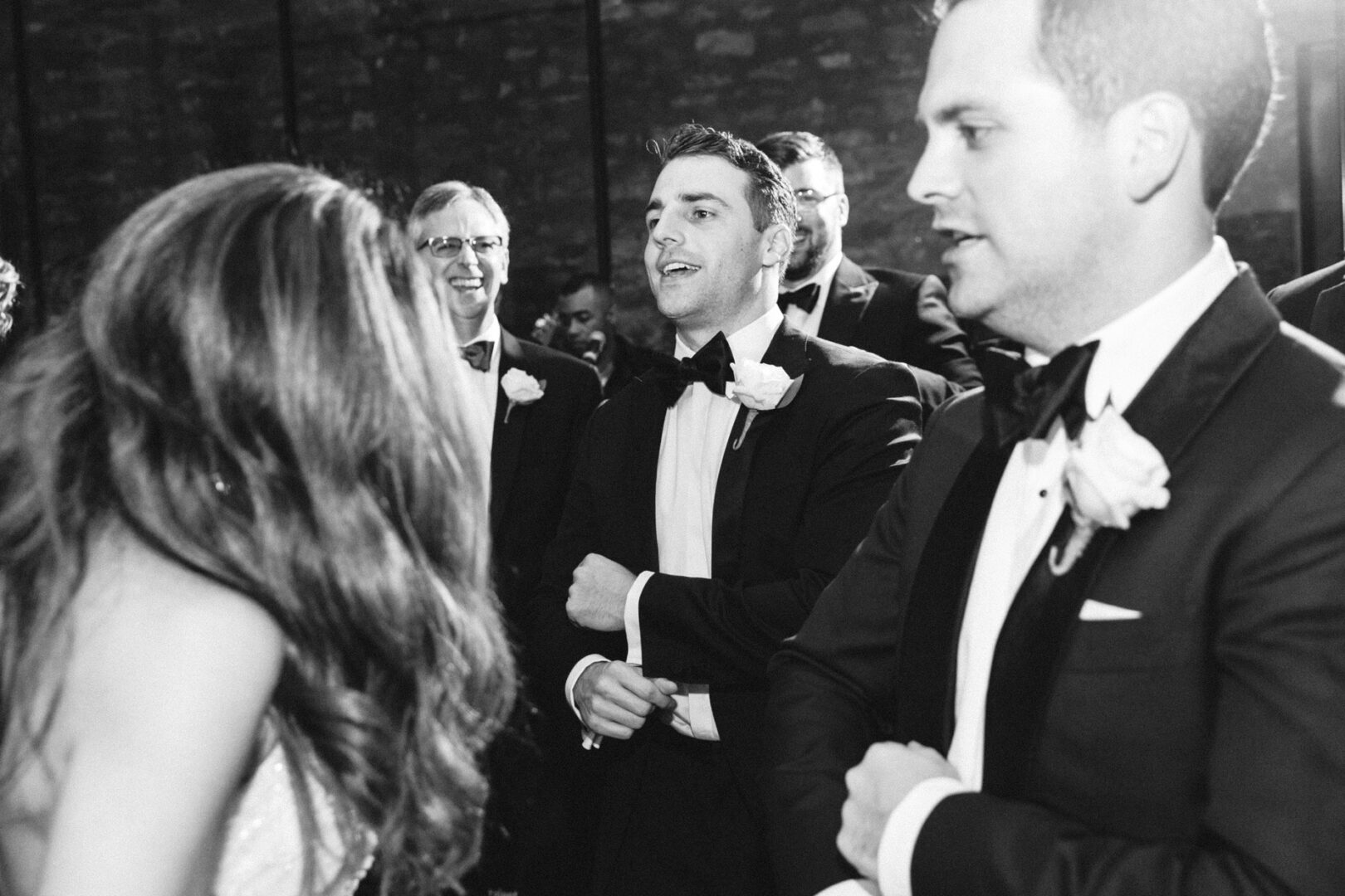 A group of people dressed in formal attire, including tuxedos, mingles elegantly indoors at the Michener Art Museum. Some guests turn their attention toward a woman in the foreground, adding an air of sophistication to the wedding gathering.