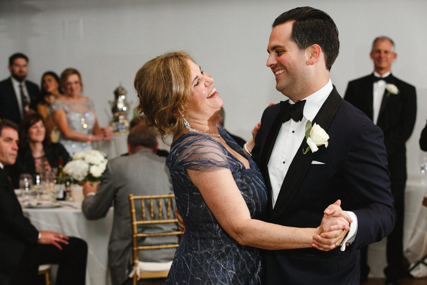 A man and woman in formal attire dance joyfully at a Michener Art Museum wedding celebration. Others are seated and standing in the background, dressed elegantly.