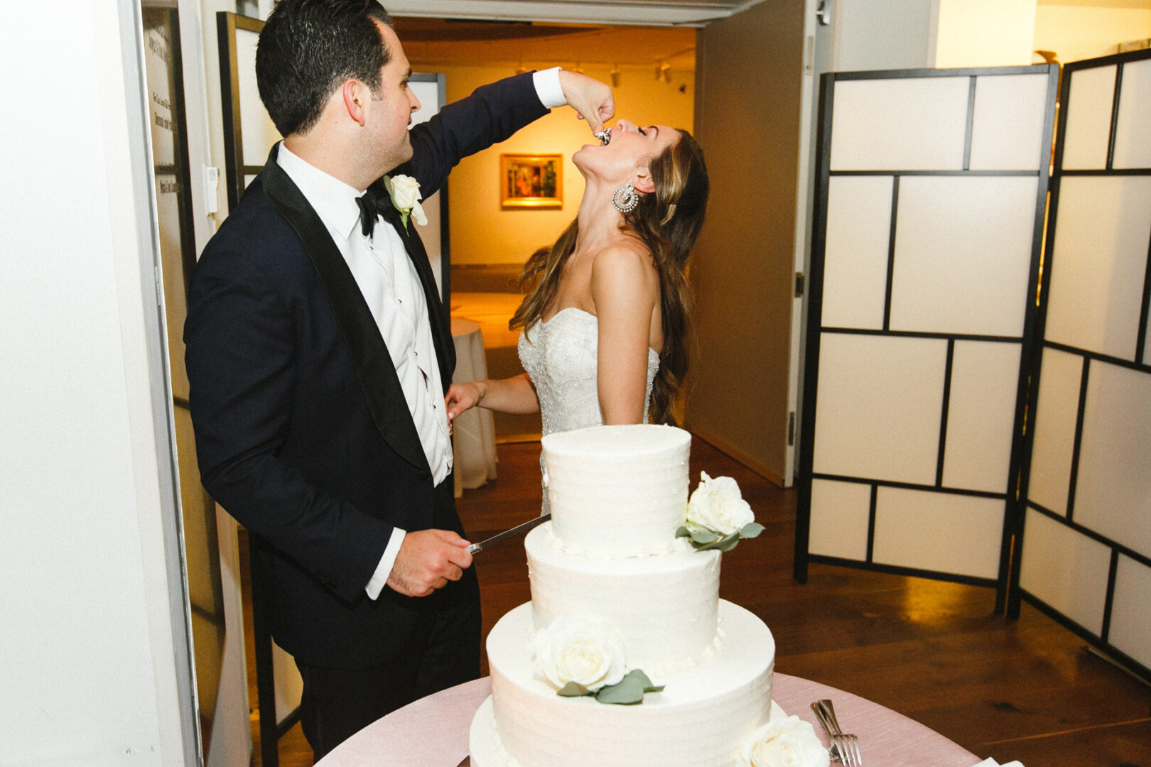 At a charming Michener Art Museum wedding, a bride and groom share a sweet moment as he playfully feeds her cake. In the foreground, a three-tiered masterpiece adorned with roses complements the joy of the day.