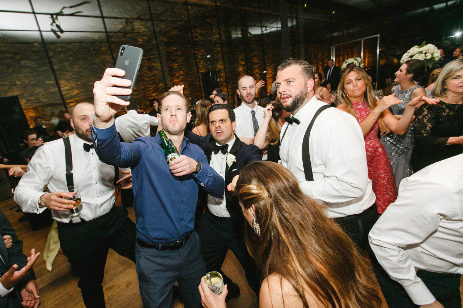 A group of people in formal attire gather closely, some holding drinks, at what appears to be a Michener Art Museum wedding. One person in a blue shirt takes a selfie against the backdrop of a dimly lit room with an elegant stone wall.