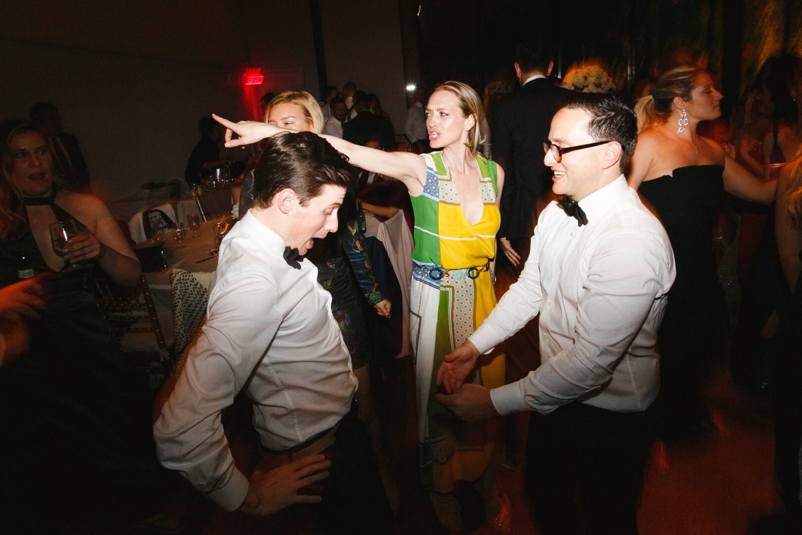 People in formal attire are dancing and interacting at a lively indoor event, resembling a Michener Art Museum wedding, with one woman pointing.