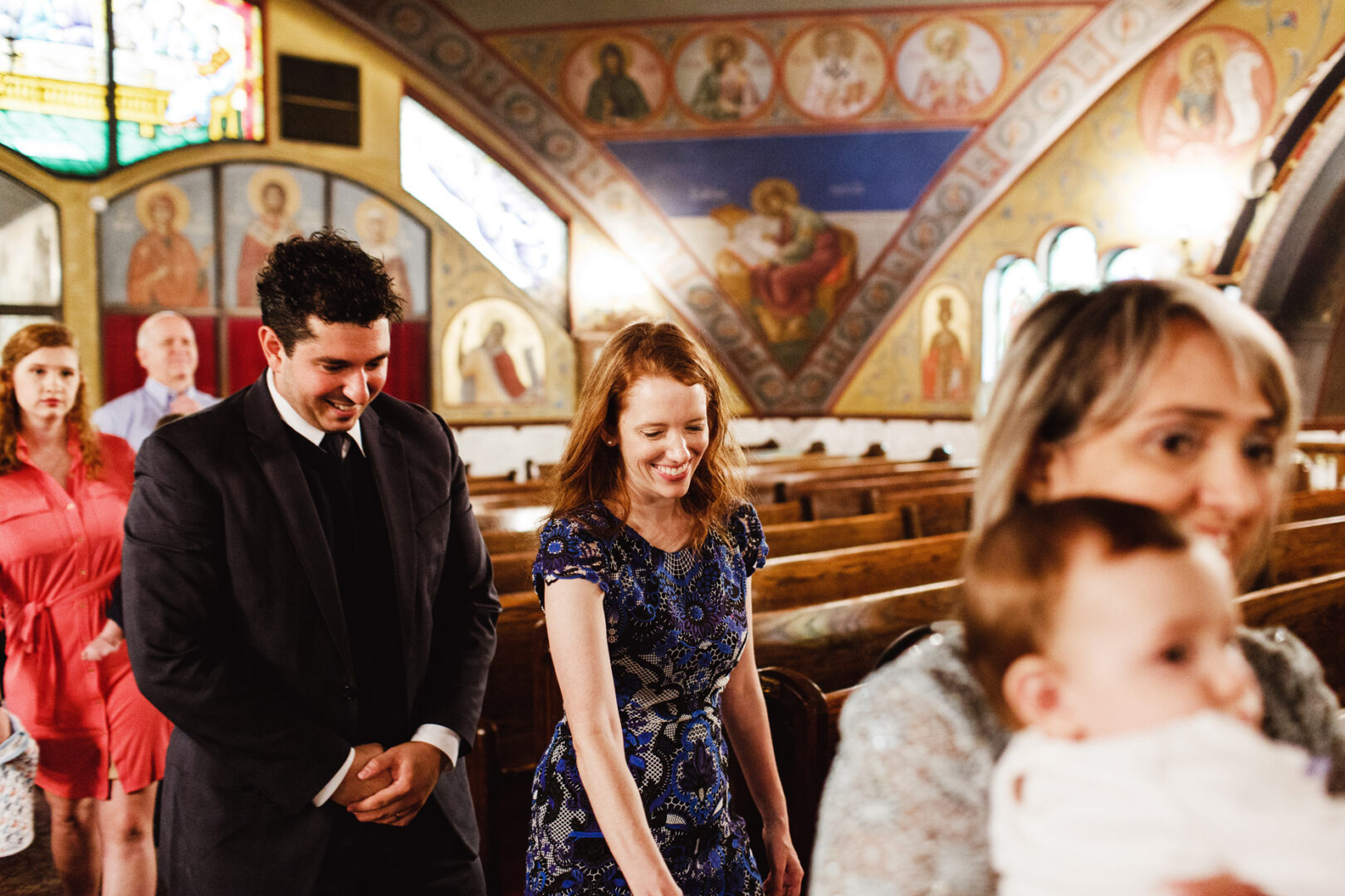 A group of people, including a woman cradling a baby, walks down the aisle of a church adorned with religious art, capturing the essence of New York City baptism photos.
