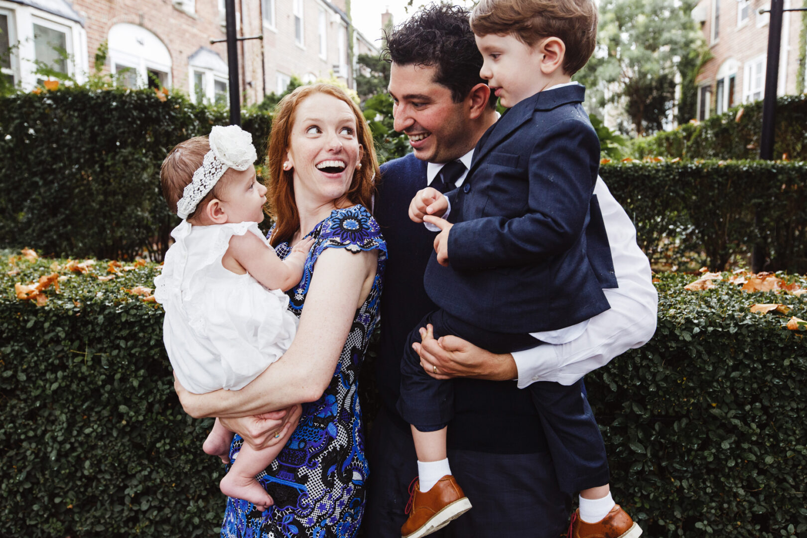 A couple stands outdoors amidst lush greenery, each holding a child. The woman cradles a baby in a white outfit and headband, while the man holds a toddler in a blue suit. They are all smiles, capturing the joy of their New York City baptism photos.