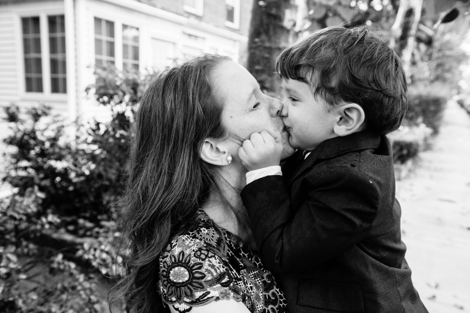 A woman holds a young boy close, their noses touching, both smiling outdoors in a residential area. The black and white photo captures a serene moment reminiscent of cherished New York City Baptism Photos.