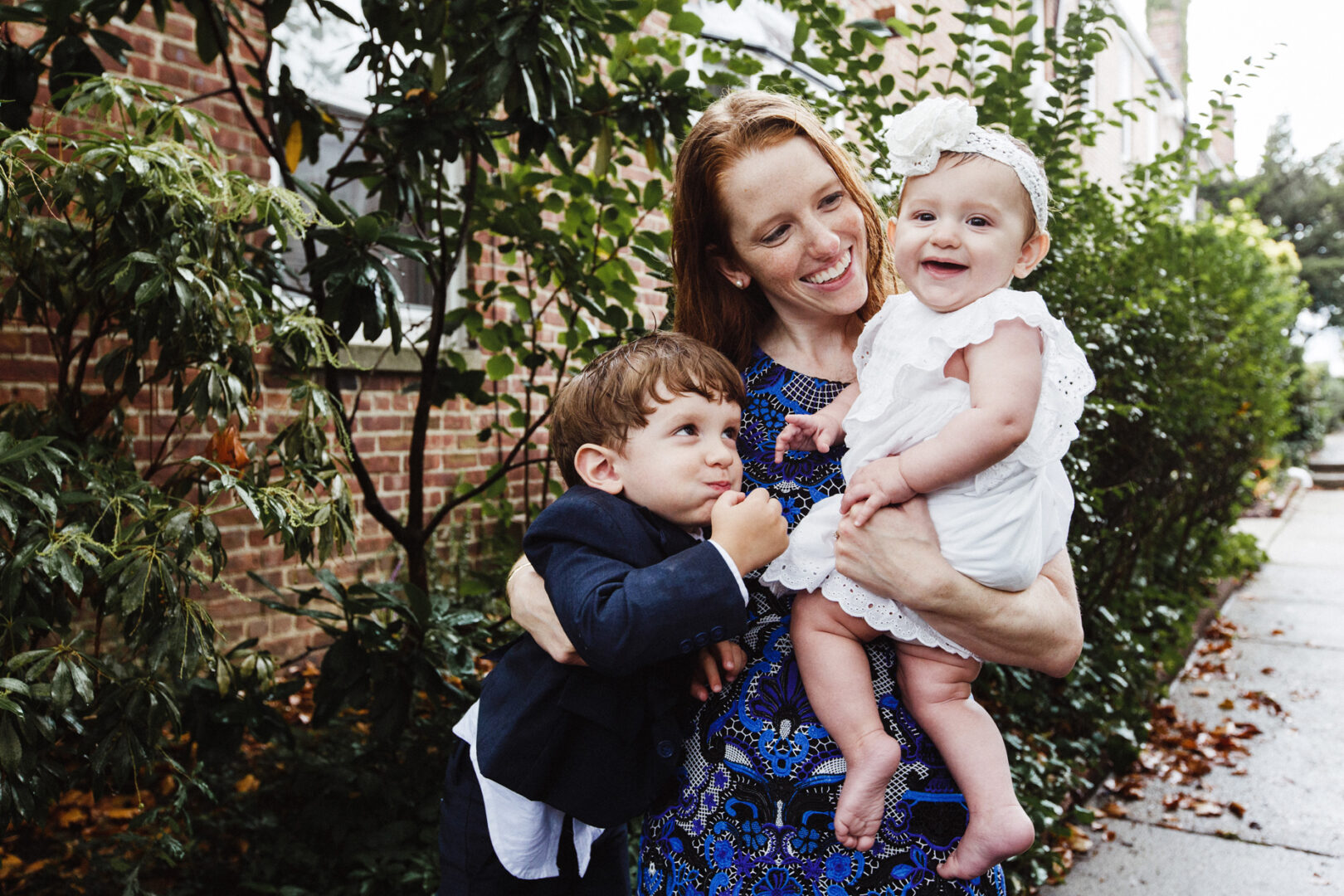 A woman smiles as she holds a baby in white, celebrating a New York City baptism. She stands beside a young boy in a suit, surrounded by green plants and a brick wall, capturing the joy of the day.