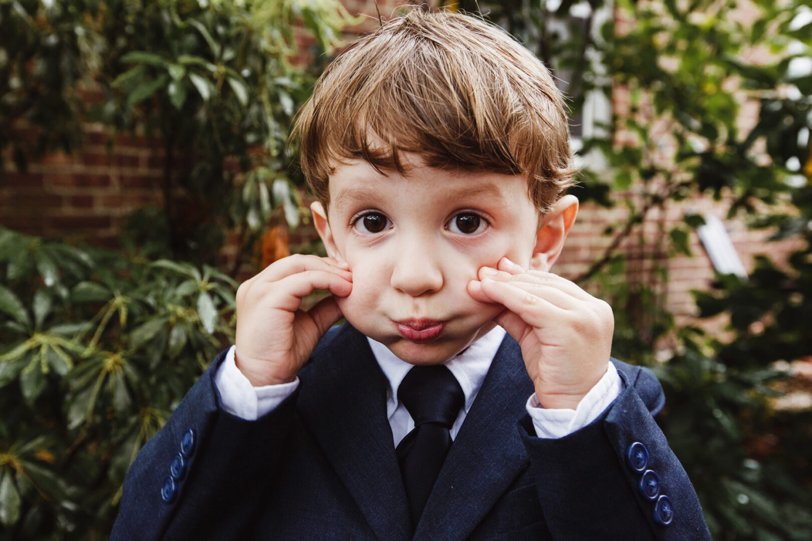 A young boy in a suit, exuding charm, playfully pulls his cheeks against the backdrop of lush green foliage and a brick wall—a delightful moment captured in New York City baptism photos.