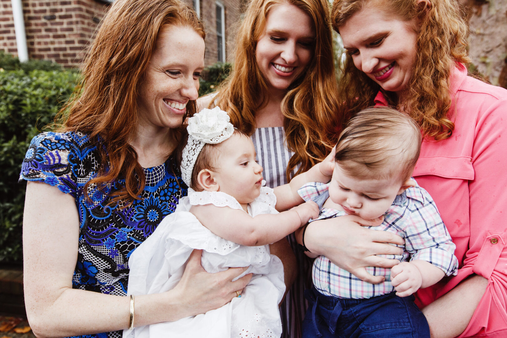 Two women holding babies stand closely together, smiling. Another woman stands between them, adoring the infants. Captured near a brick building in New York City, this charming scene is part of a cherished baptism photo collection.