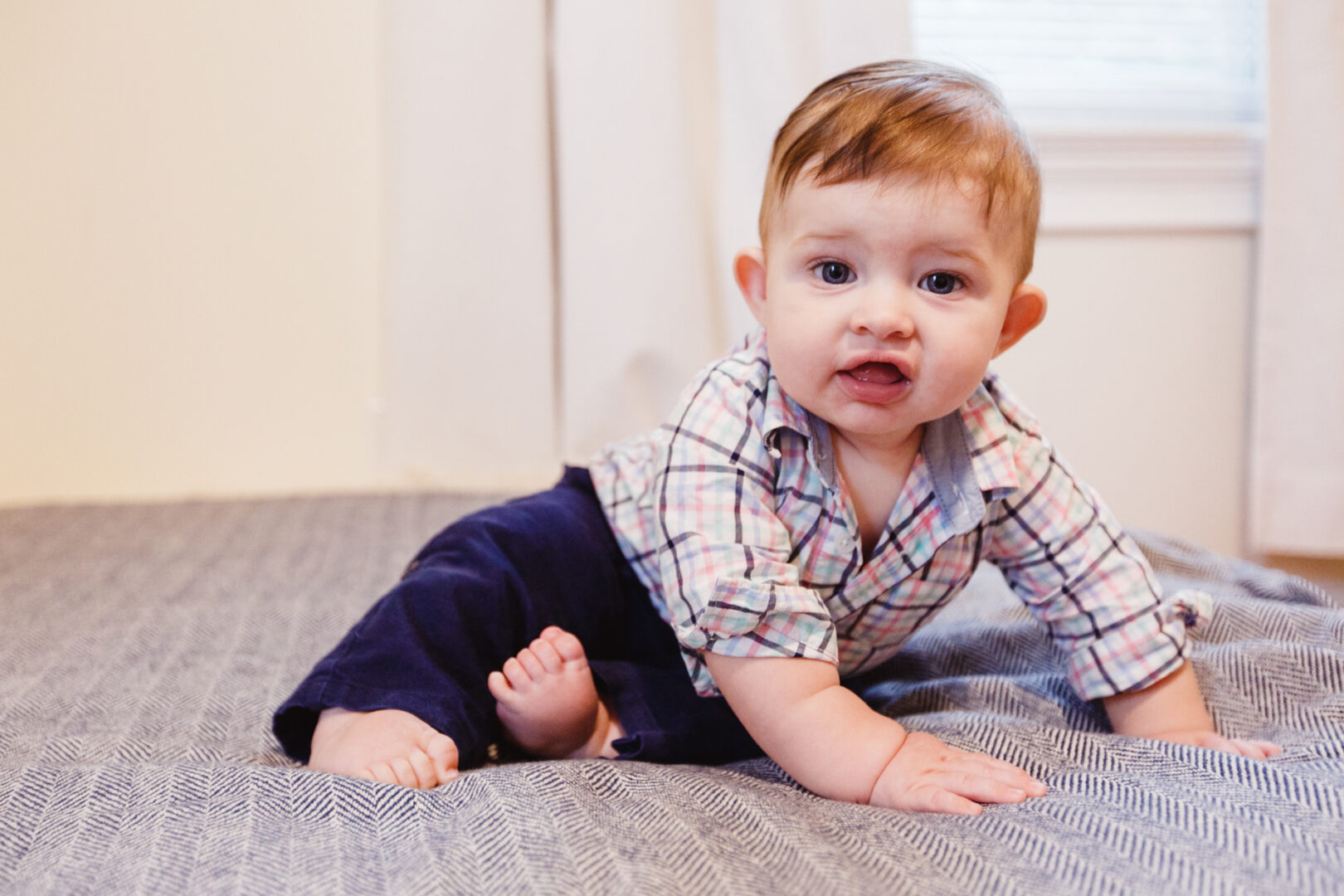 A baby in a plaid shirt and dark pants crawls on a bed with a neutral-colored blanket, near a window with white curtains, ready for their New York City baptism photos.