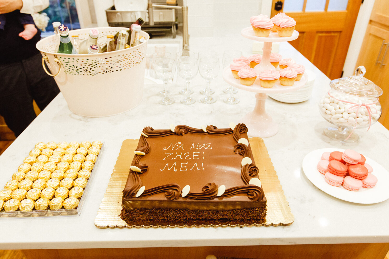 A table adorned with a large chocolate cake featuring Greek writing, surrounded by macarons, cupcakes, chocolates, a drink basket, and a jar of mints. Perfect for capturing memorable moments in New York City baptism photos.