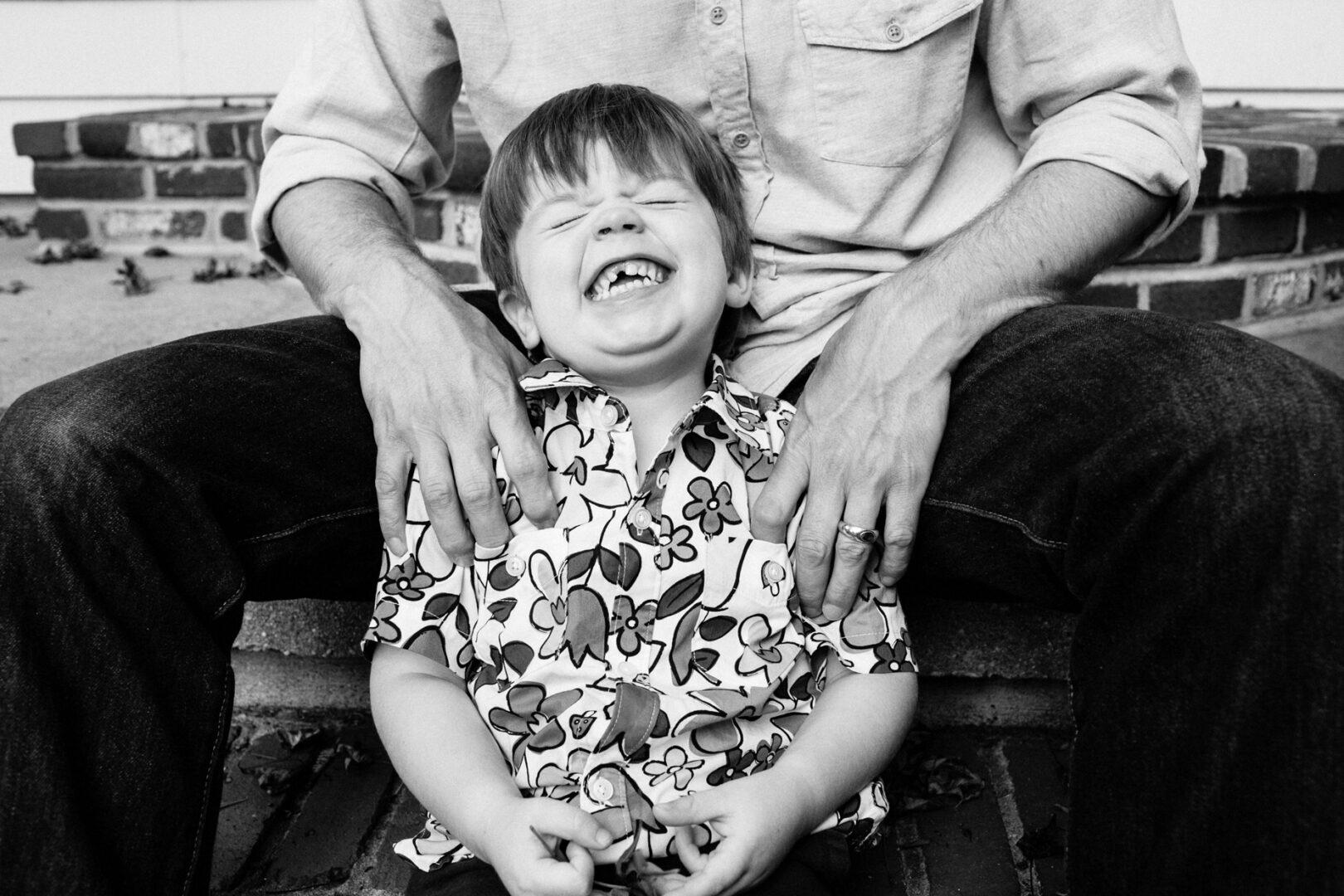 A child sits on an adult's lap, smiling widely with eyes closed in a heartwarming Philadelphia family photo. The child wears a floral shirt while the adult sports jeans and a casual top. Behind them, brick steps create a charming backdrop.