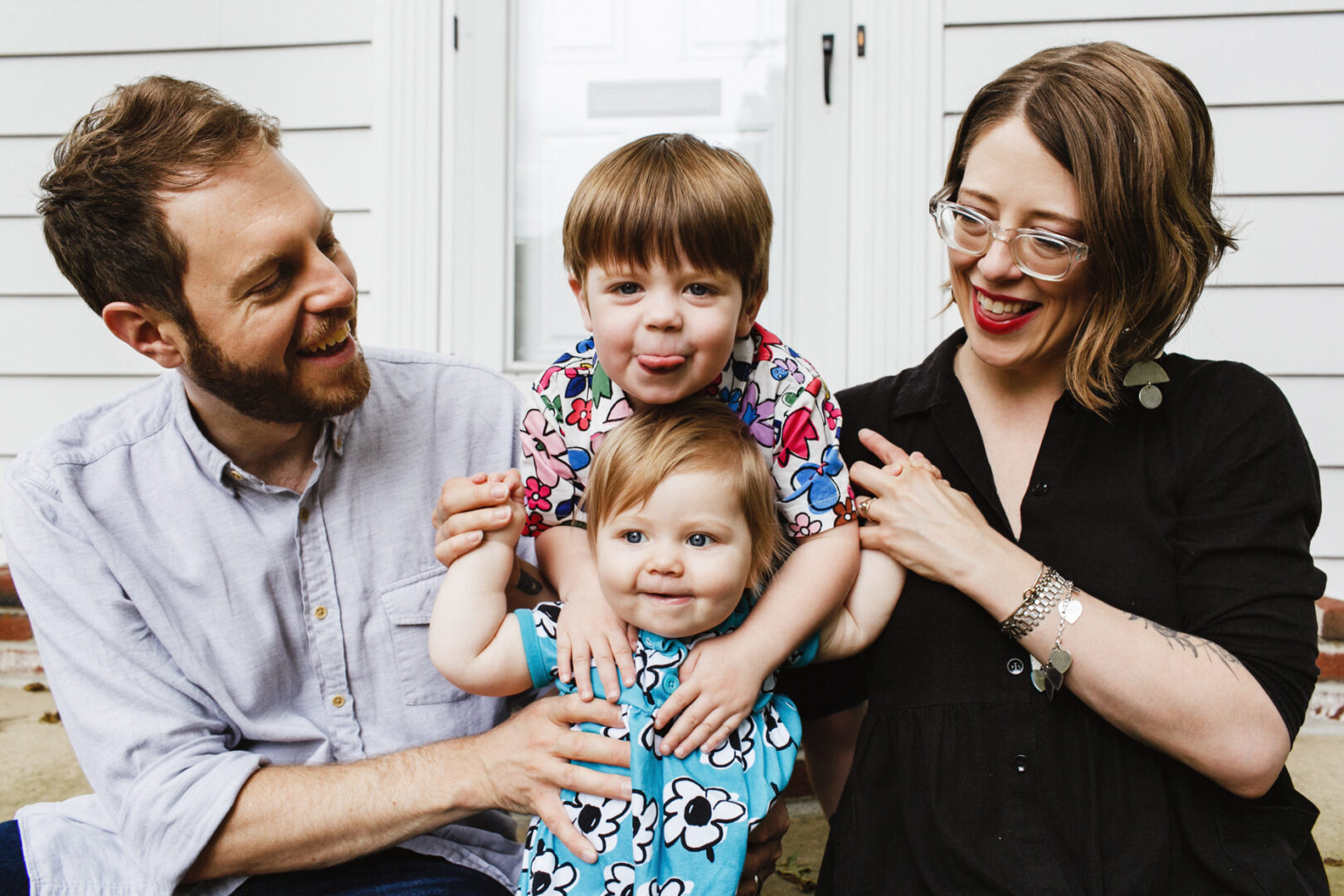 A joyful Philadelphia family gathers outside a white building, as one child playfully balances on a sibling's shoulders. They're all smiling, capturing the essence of treasured family photos filled with warmth and connection.